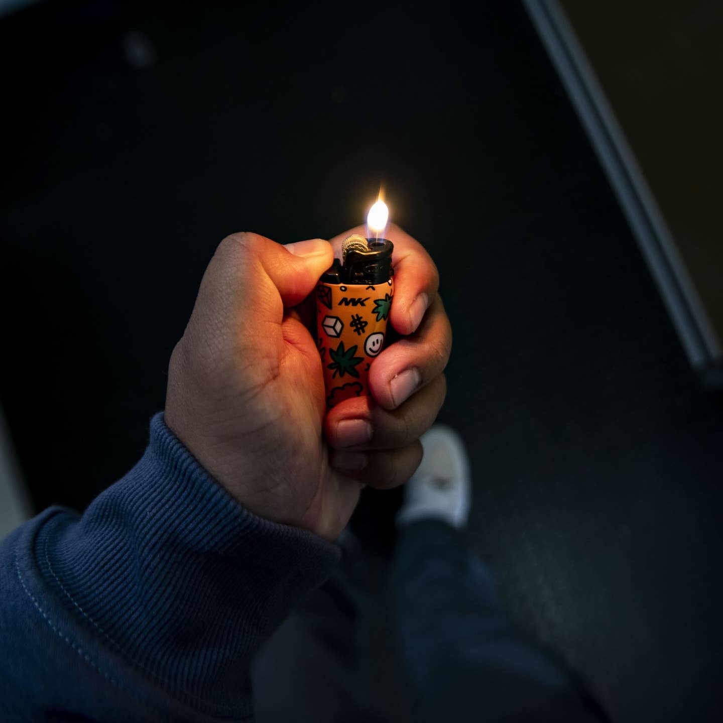A person in a blue sleeve holds an MK Lighter Company Flint Strike Refillable Lighter, featuring colorful leaf and symbol designs. Its flame and replaceable flint are clearly visible against a dark background.