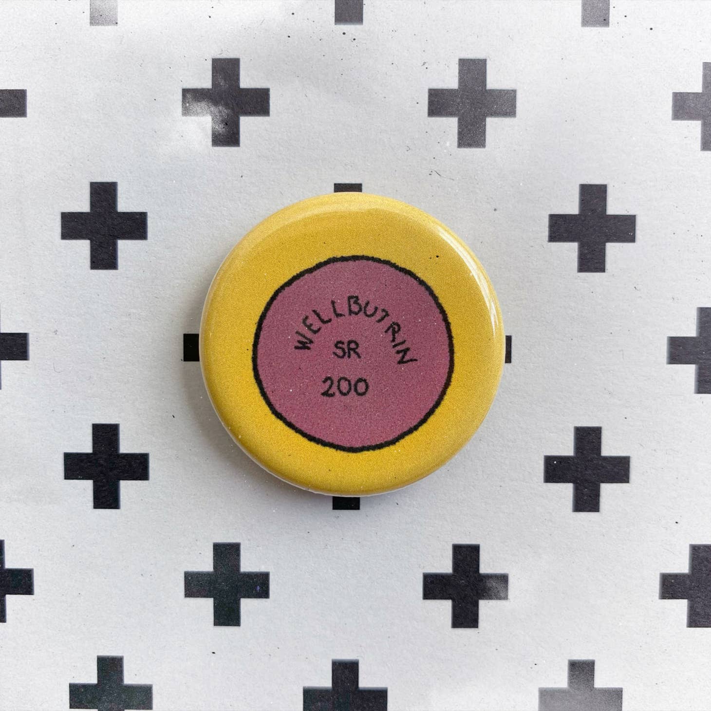 The Pill Pin by Chateau Blanche is a round accessory resembling a yellow and pink WELLBUTRIN SR 200 pill, displayed on white paper with a black plus sign pattern—an eye-catching mental health statement piece.
