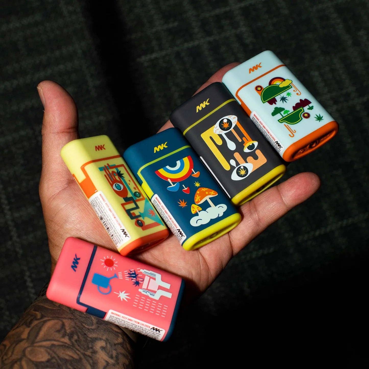 A hand holds five colorful rectangular Ignition Pocket Lighters from the MK Lighter Company’s Nature Series, each with playful cartoon designs and the letters MK on top, resting against a dark textured surface.
