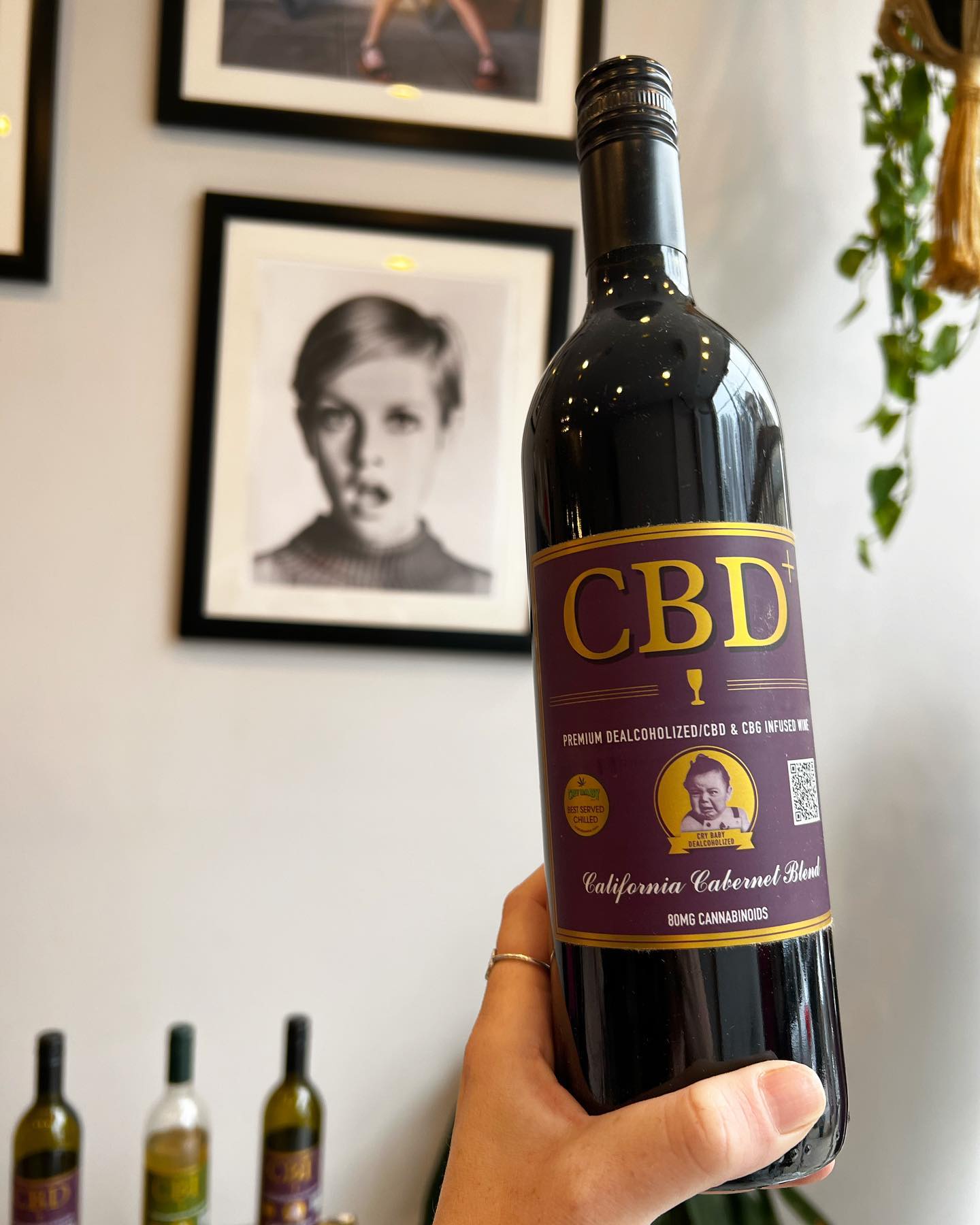 A hand holds a Cry Baby Wine “CBD Cabernet” de-alcoholized wine bottle featuring a vintage-style woman’s portrait. Black-and-white photos frame the background, and more CBD Cabernet bottles are displayed on the shelf below.