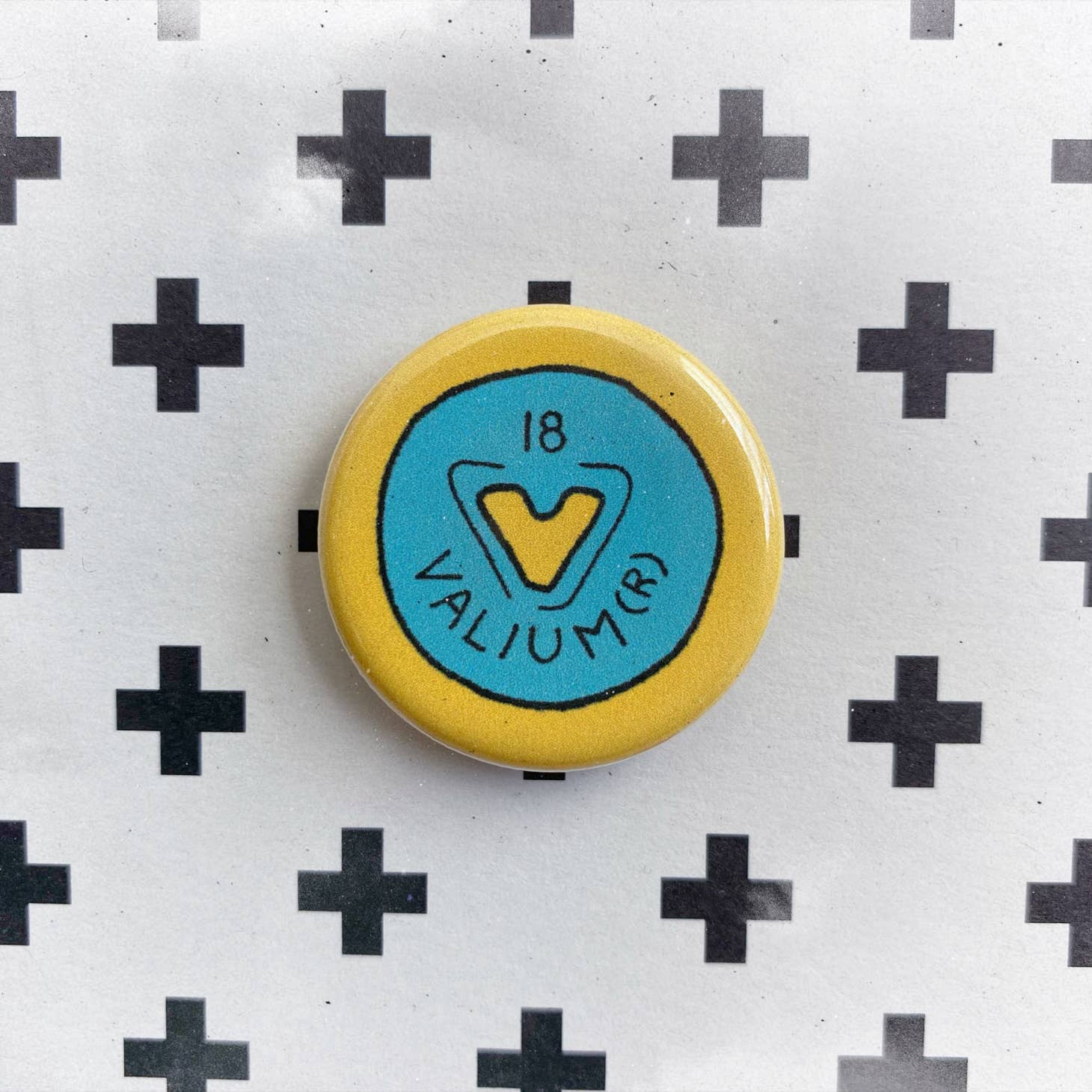 The Chateau Blanche Pill Pin, inspired by Valium with a blue center, yellow border, and 18 VALIUM (R) text, sits on a white surface with black plus signs—a standout mental health accessory.