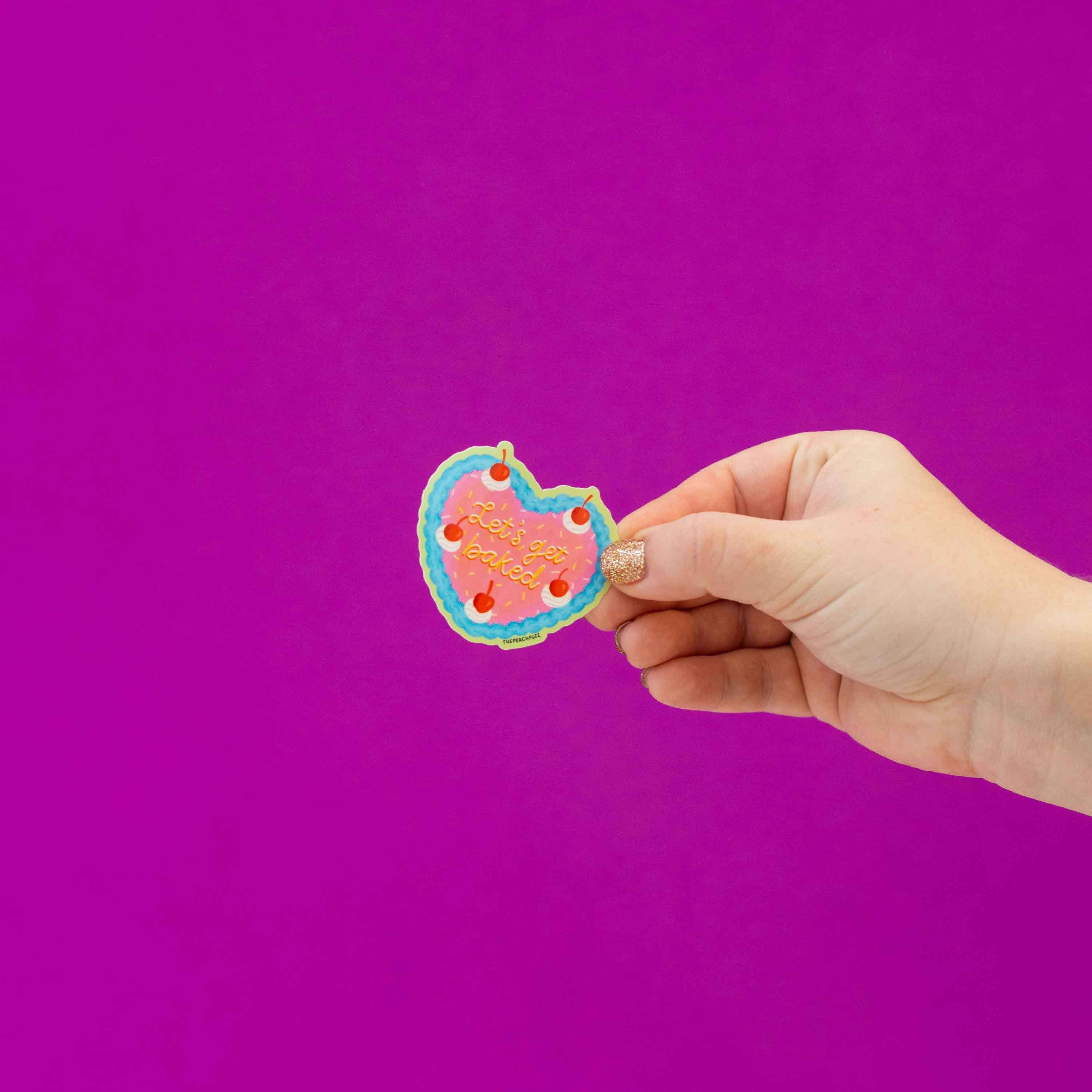 A hand with glittery nail polish holds the fun Lets Get Baked Cake Sticker by The Peach Fuzz, resembling a bitten, colorful iced cookie, set against a vibrant magenta background.