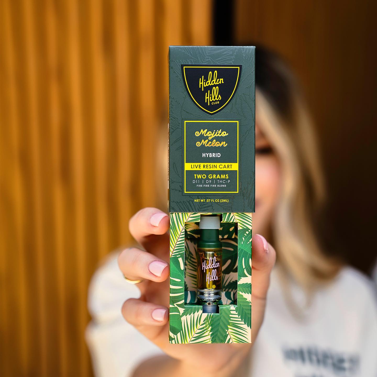 A person holds a Hidden Hills Live Resin THC Cartridge 2G box near the camera, making the packaging clear while the background with the person remains blurred.