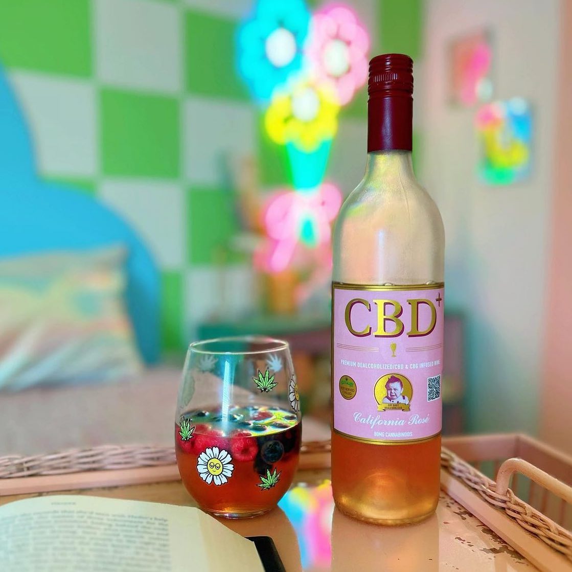 A bottle of Cry Baby Wine CBD Rosè sits beside a glass decorated with berries and marijuana leaves on a tray with an open book. Neon flower lights and a checkered wall create a colorful, blurred background.