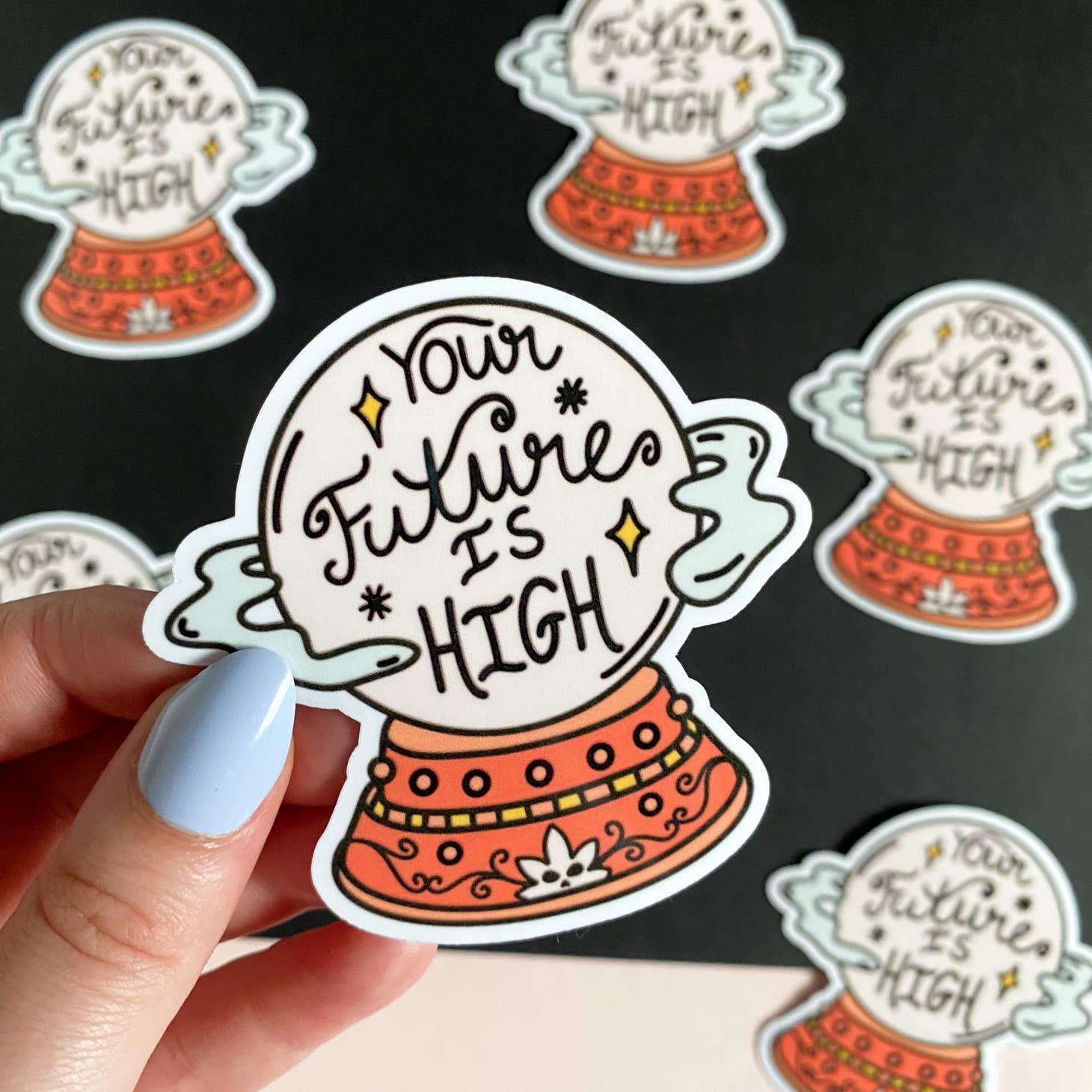 A hand with light blue nail polish holds the Han Made Your Future Is High Sticker, a waterproof design ideal for laptops, shown among similar stickers on a black surface.