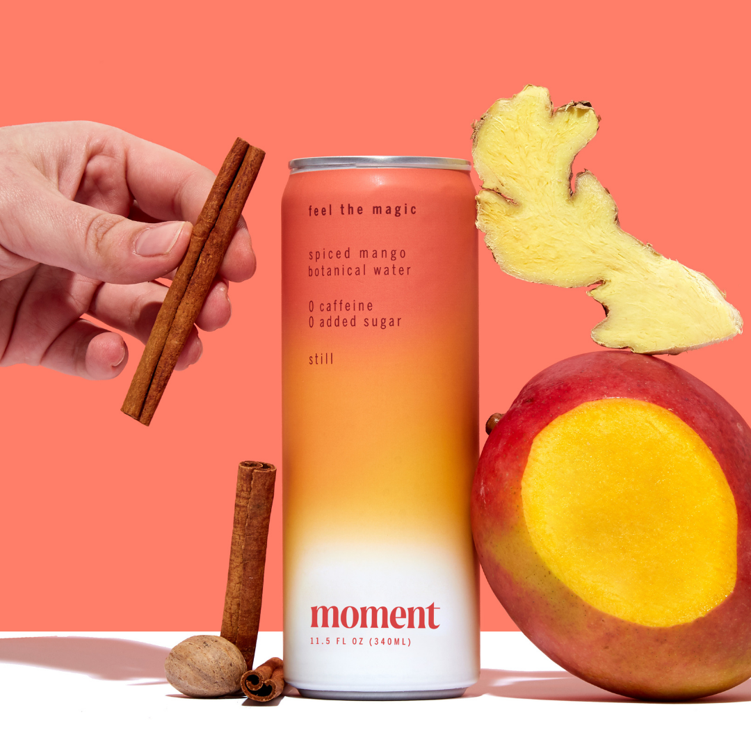 A hand holds a cinnamon stick above a can of Moment Botanical Beverage by Moment, surrounded by mango, ginger, nutmeg, and another cinnamon stick, all set against an orange background.