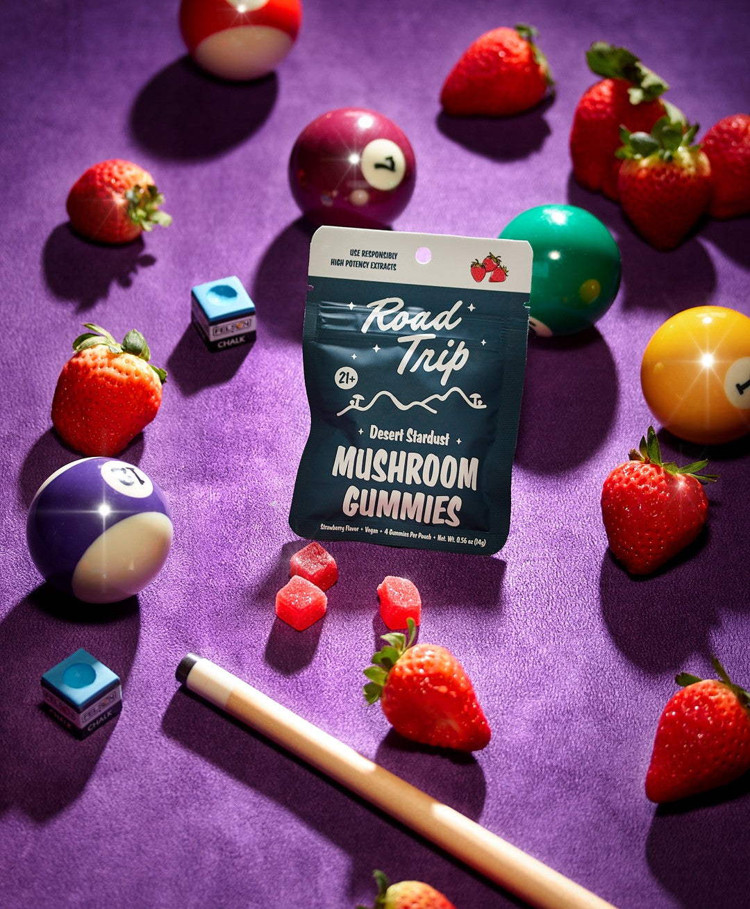 A bag of Road Trip Mushroom Gummies by Road Trip sits on a purple pool table surrounded by billiard balls, strawberries, a pool cue, chalk, and several loose gummies.