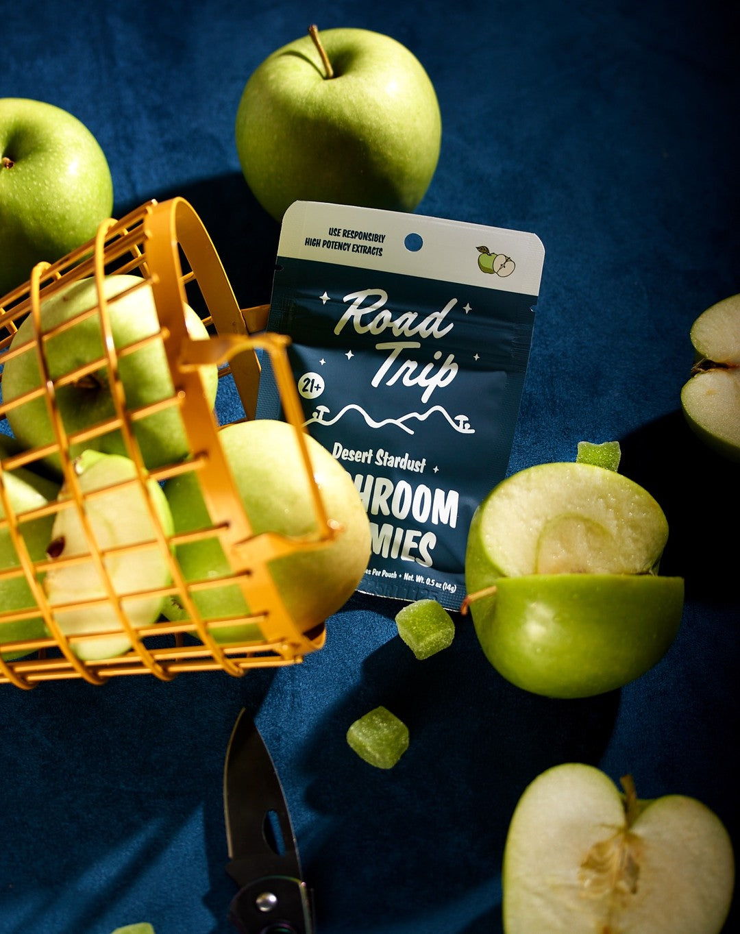 A yellow basket of green apples spills onto a blue surface near a package of Road Trip Mushroom Gummies by Road Trip, with sliced green apples and small plant-based gummies scattered around. Scissors are visible at the bottom of the image.