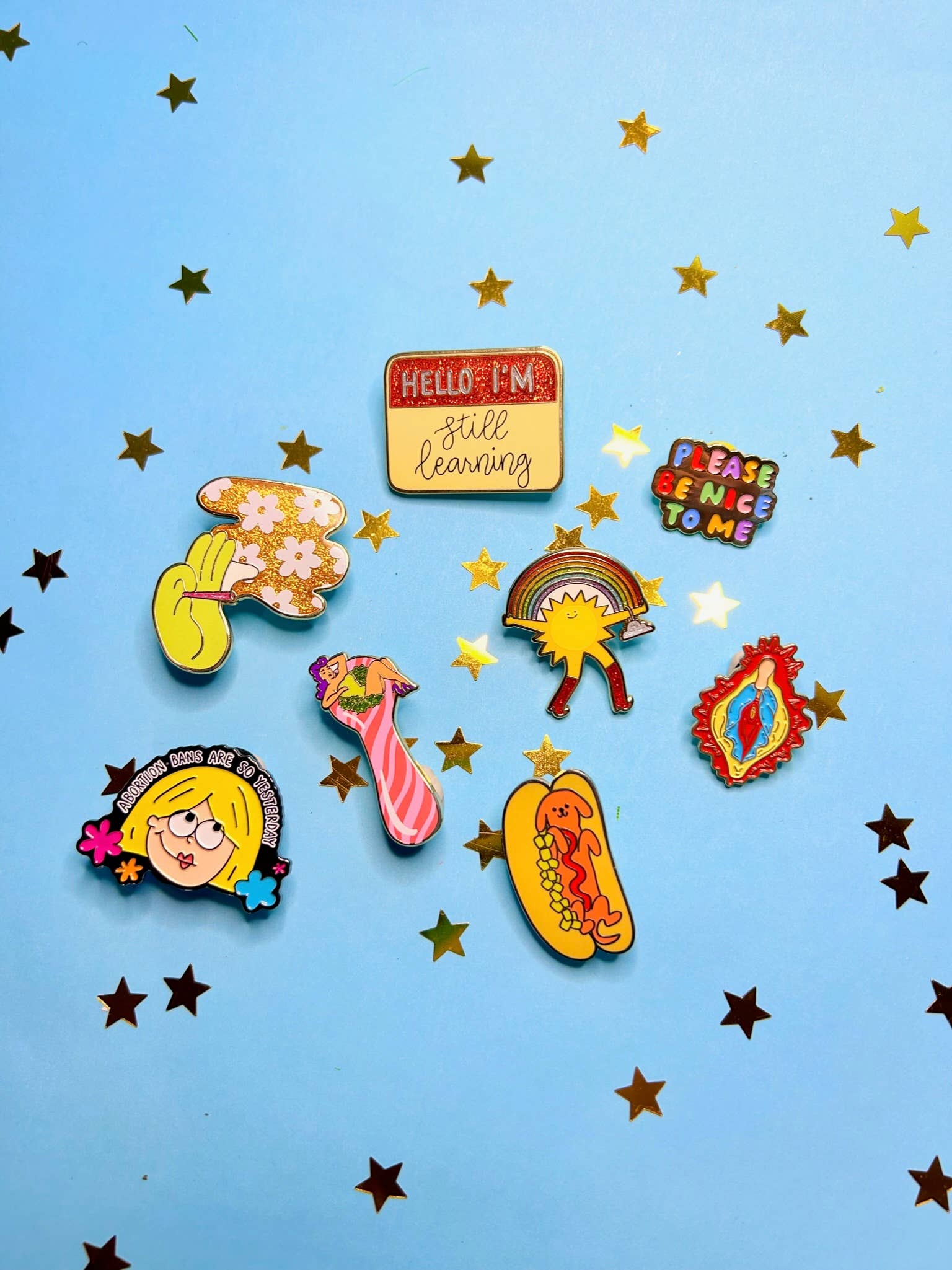 The Peach Fuzzs Total Smoke Show Enamel Pin shines among other colorful enamel pins, including a glittery design and an uplifting pinup girl pin, all set on a light blue background with gold star confetti.