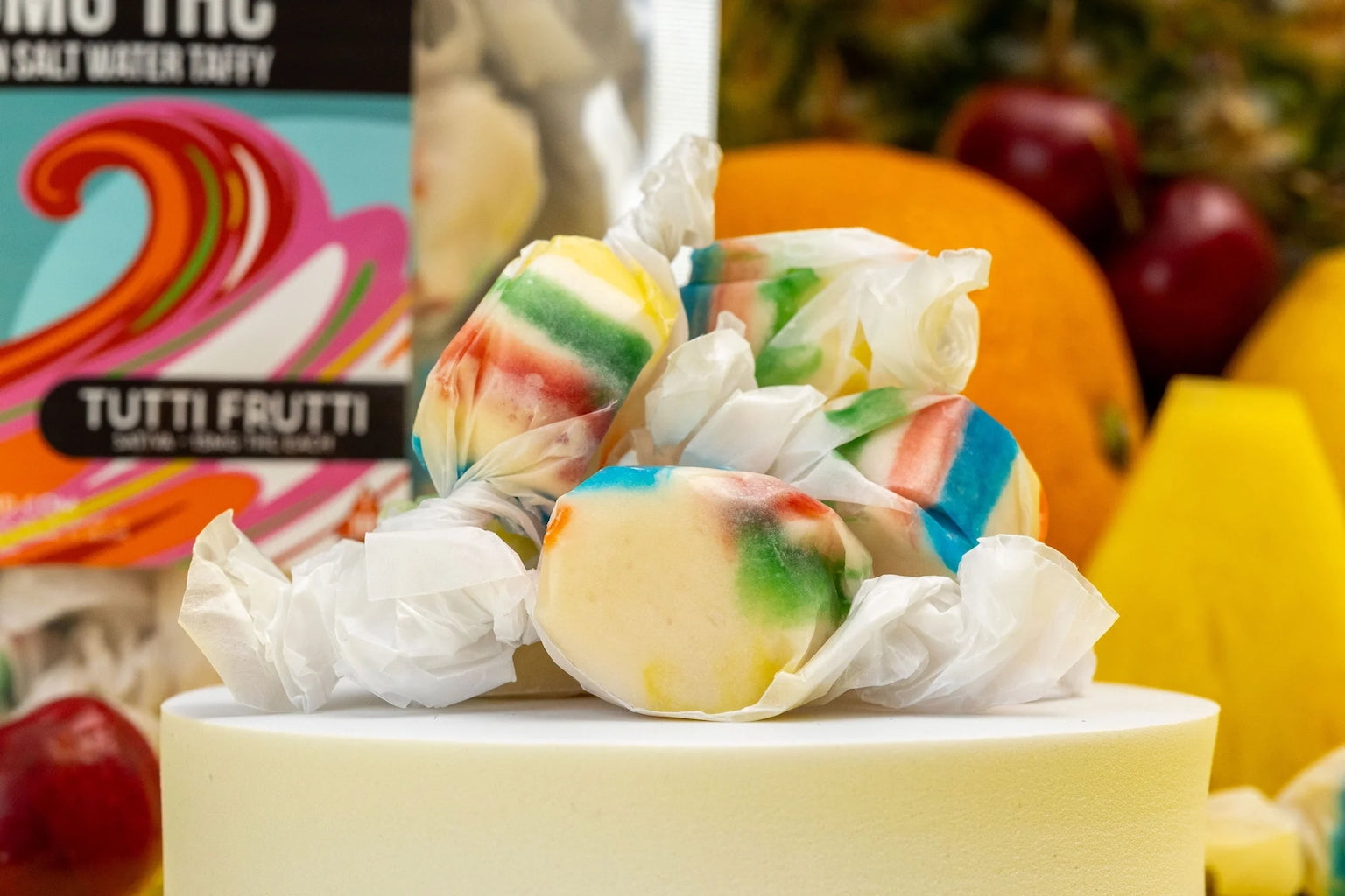 A stack of Hometown Hero Delta-9 Live Rosin Taffy, wrapped in white paper, sits on a white surface with colorful fruits and a taffy box blurred in the background.