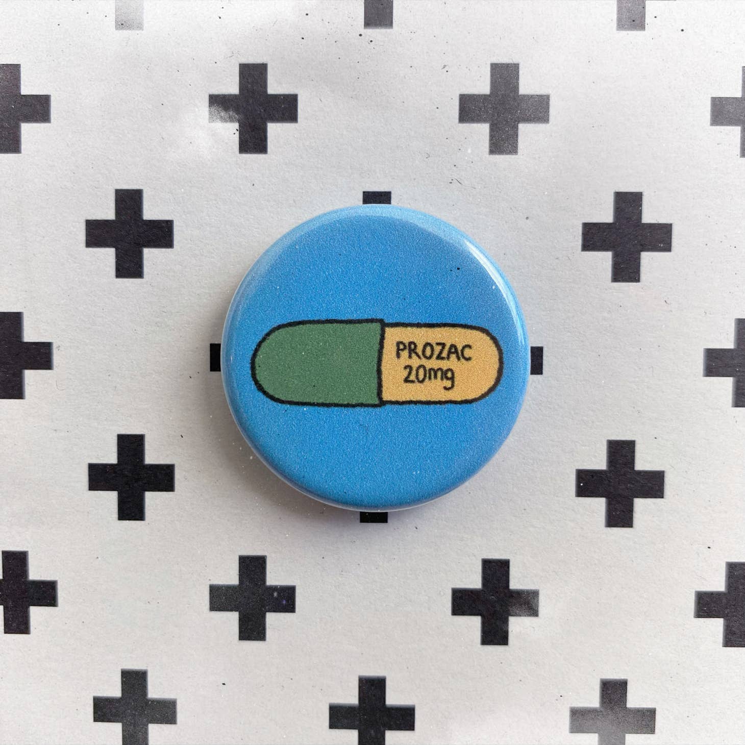The Chateau Blanche Pill Pin is a round blue button with a green and yellow PROZAC 20mg capsule illustration on a black plus sign pattern—an eye-catching and unique mental health accessory.