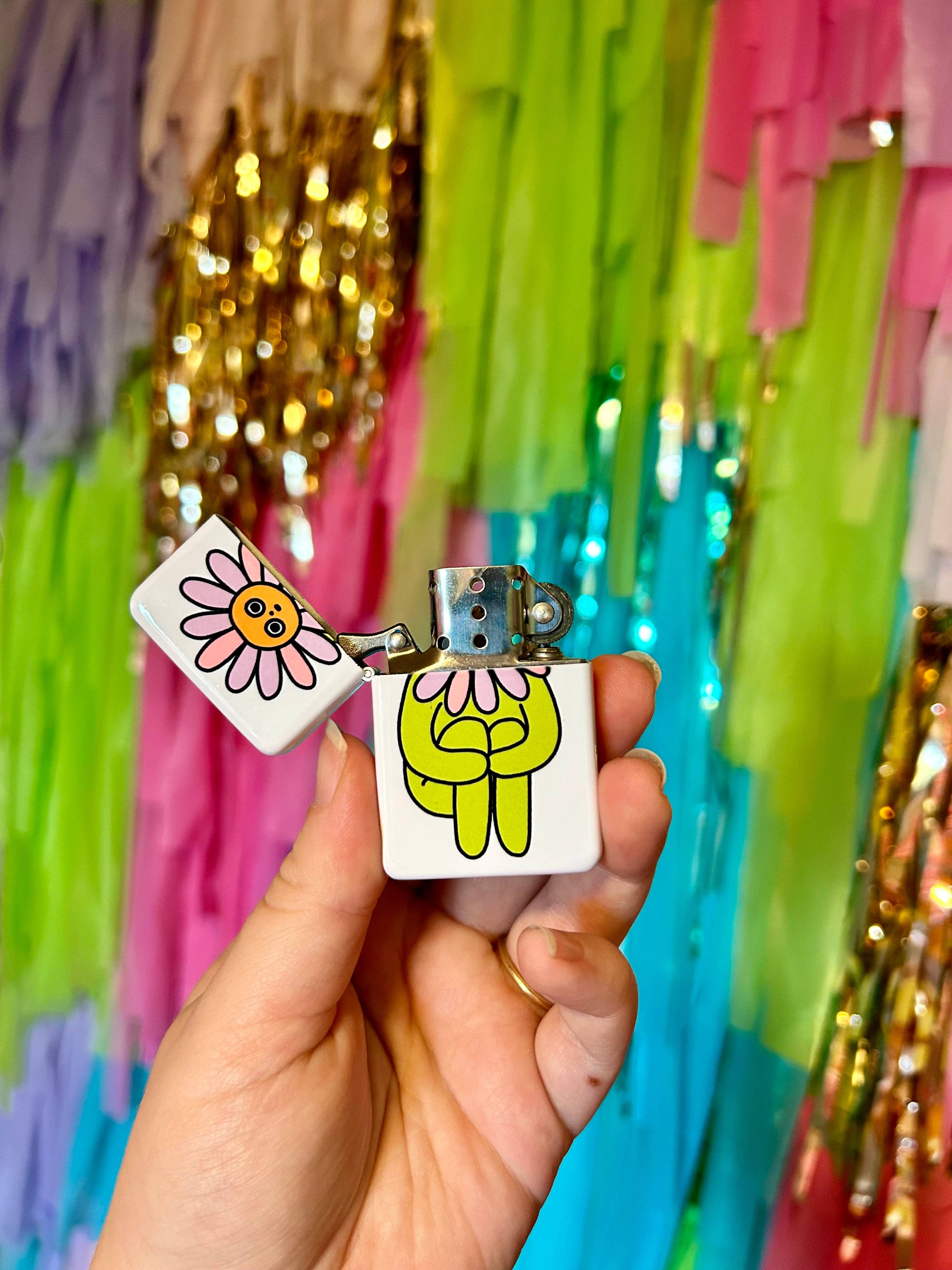 A hand holds The Peach Fuzz’s Flower Boy Lighter: Perfect, a refillable lighter featuring a cute flower design—ideal for candle lovers—set against pastel and gold fringe decorations.