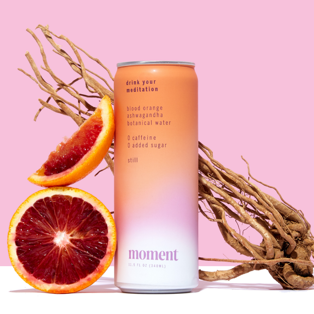 A halved blood orange and slice rest beside a pastel gradient can of Moment Botanical Beverage, an adaptogenic drink by Moment, set against a pink background with decorative tangled roots in the back.