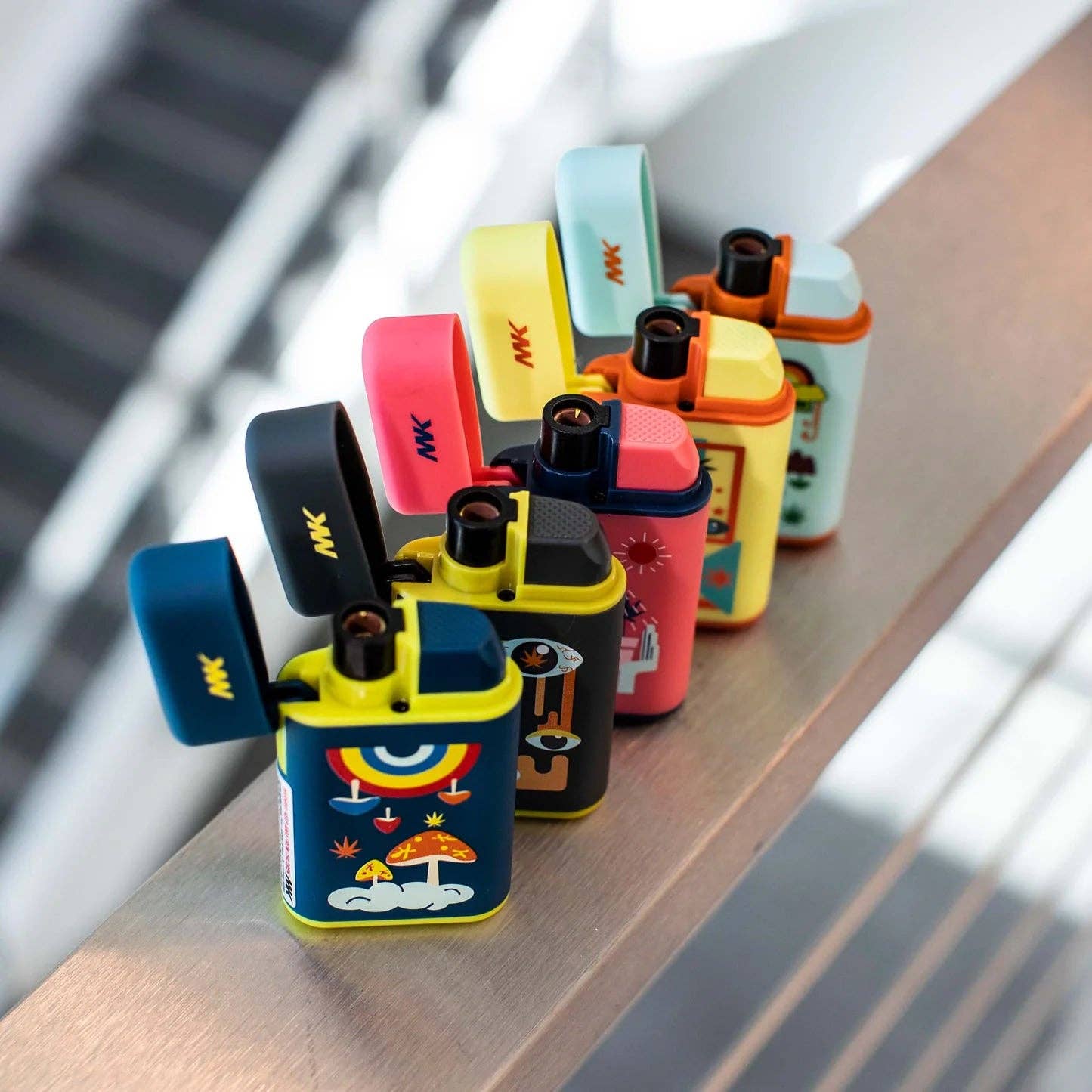 A row of vibrant Ignition Pocket Lighters - Nature Series by MK Lighter Company with playful, cartoon-like designs sits on a ledge, the blurred background highlighting the colorful refillable butane lighters in the foreground.