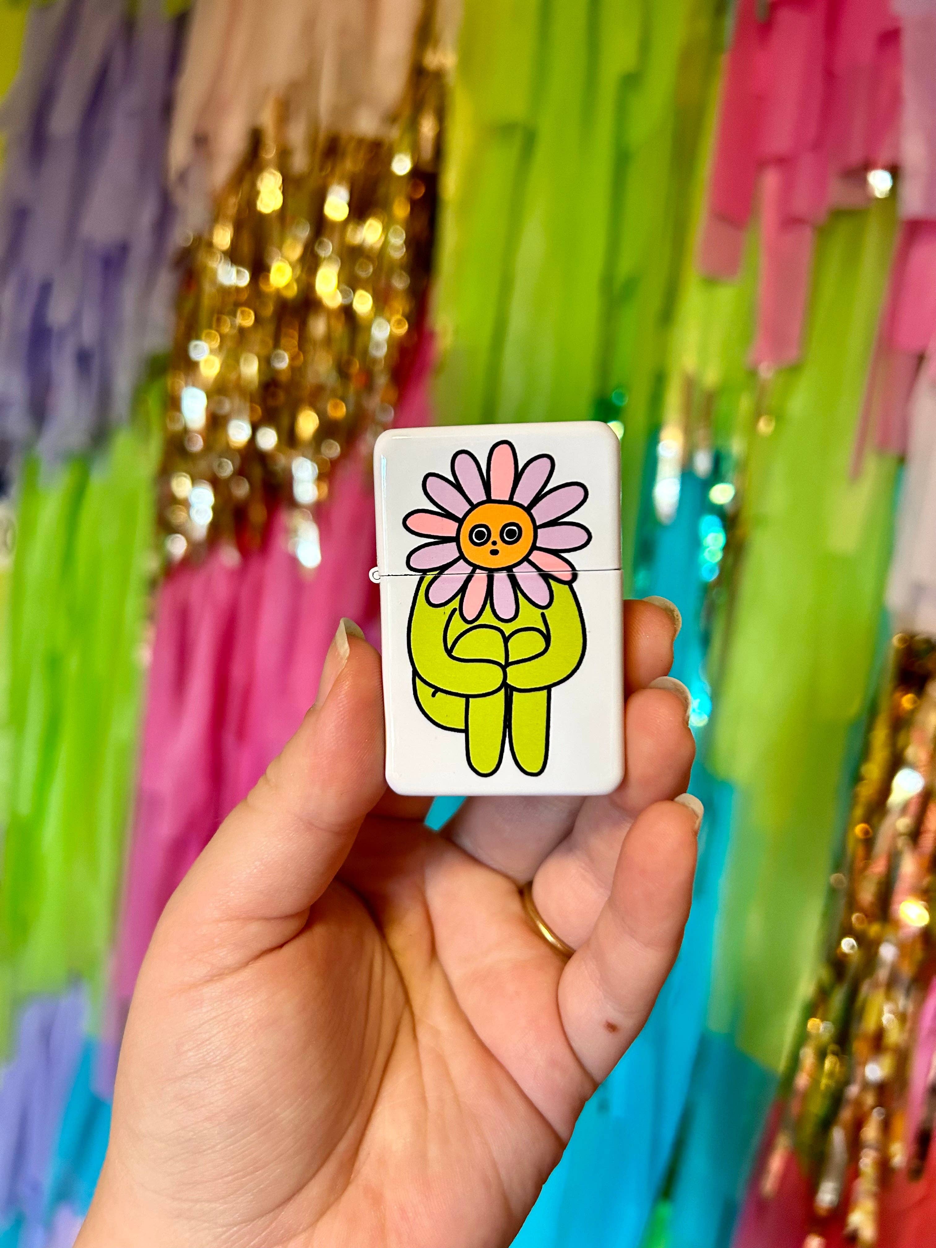 A hand holds The Peach Fuzzs Flower Boy Lighter: Perfect, a white refillable lighter with a cartoon yellow figure and pink flower head—set against a vibrant, fringed background. Ideal for stoners or candle lovers.