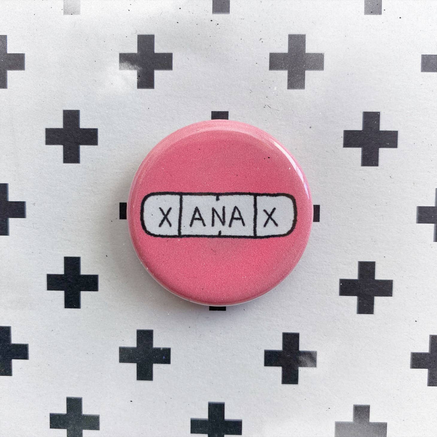 The Chateau Blanche Pill Pin is a pink accessory featuring a white rectangle with X ANA X in black letters, resembling medication, set against a background of black plus signs—a unique mental health statement piece.