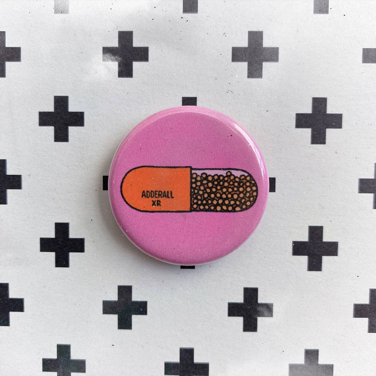 The Chateau Blanche Pill Pin is a round pink accessory featuring an Adderall XR capsule illustration on a white background with black plus signs, adding a playful touch to your mental health collection.