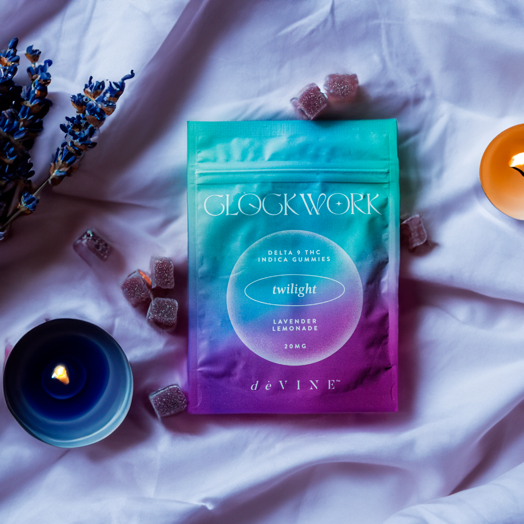 A colorful bag of DeVINE Wellness Clockwork- Twilight Indica ∆9 Gummies rests on white fabric, surrounded by candles, lavender sprigs, and scattered gummies.