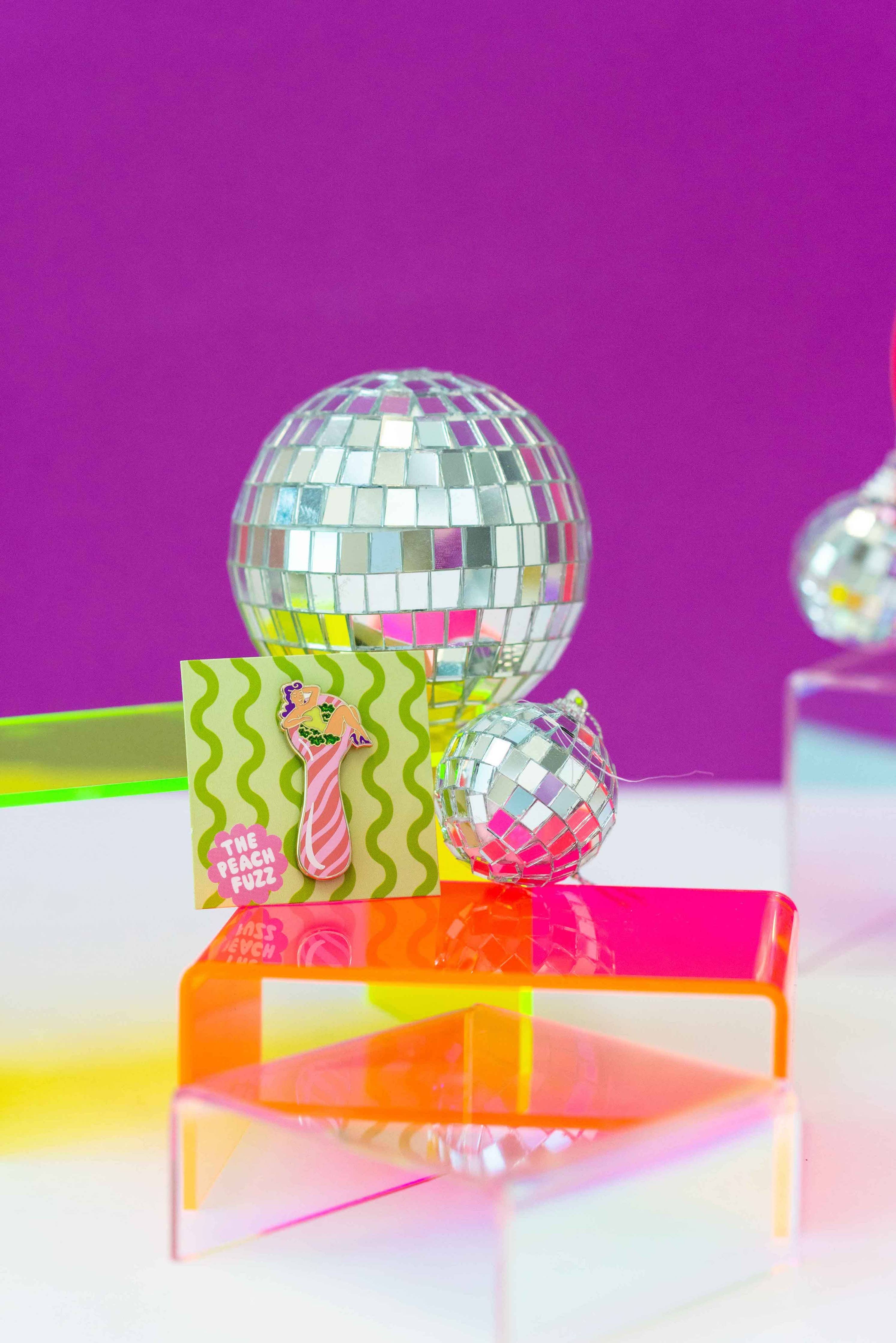 A small greeting card with a peach milkshake illustration stands beside two disco balls and The Peach Fuzz’s Total Smoke Show Enamel Pin, all displayed on colorful risers against a vibrant purple background.