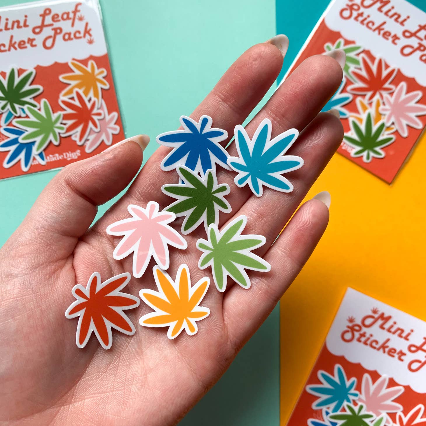 A hand displays six colorful weed leaf stickers from the Han Made Mini Rainbow Weed Leaf Sticker Pack, with packaged mini vinyl sticker packs visible on a vibrant surface in the background.
