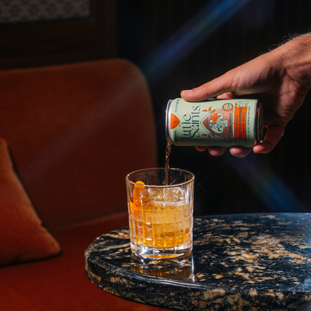 A hand pours a can of Little Saints Spiced Old Fashioned Adaptogenic Mocktail into a glass with ice and an orange peel on a dark marble table, with a brown chair and warm lighting in the background.