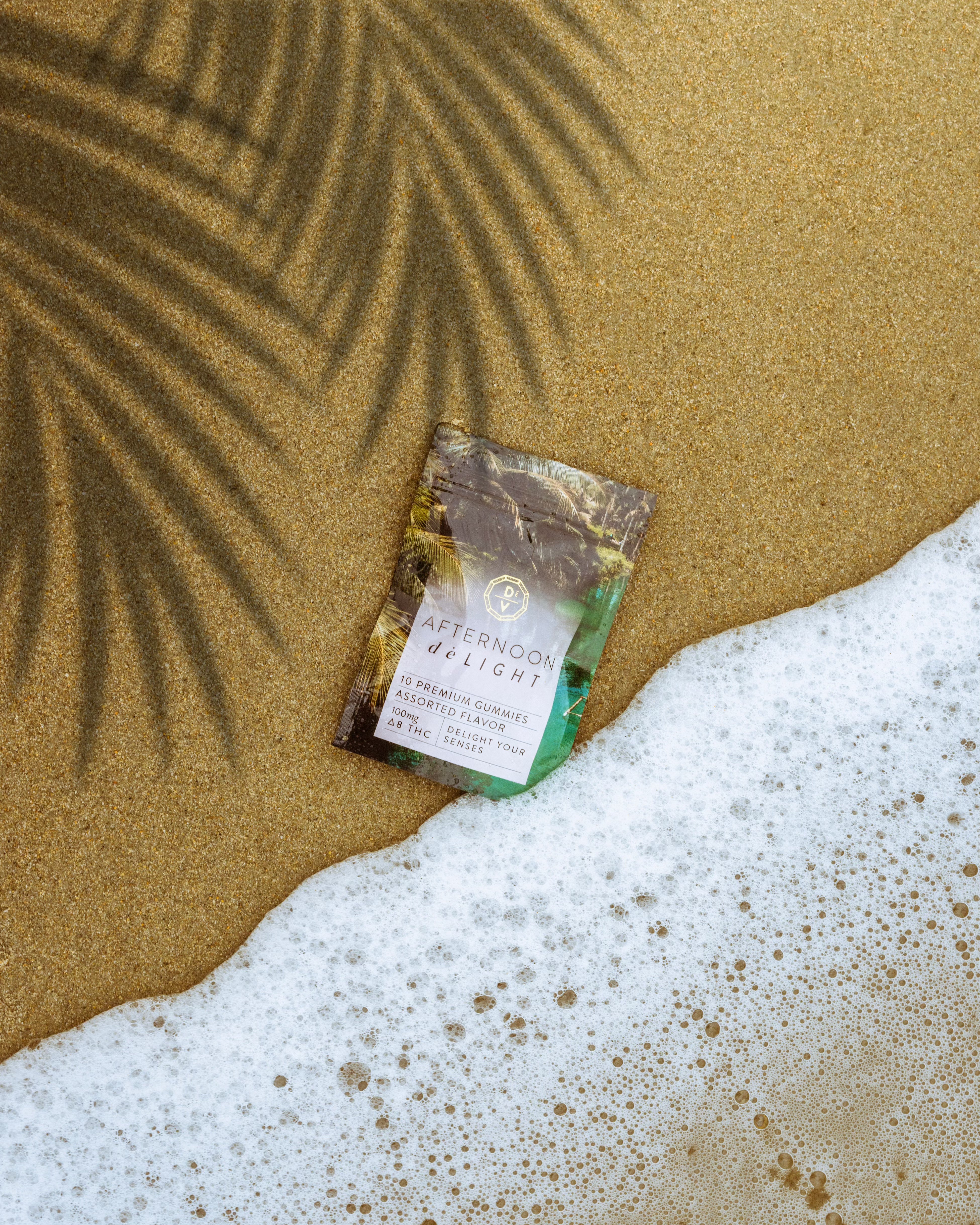 Afternoon deLIGHT ∆8 Gummies package on sandy beach near water with palm shadow in background