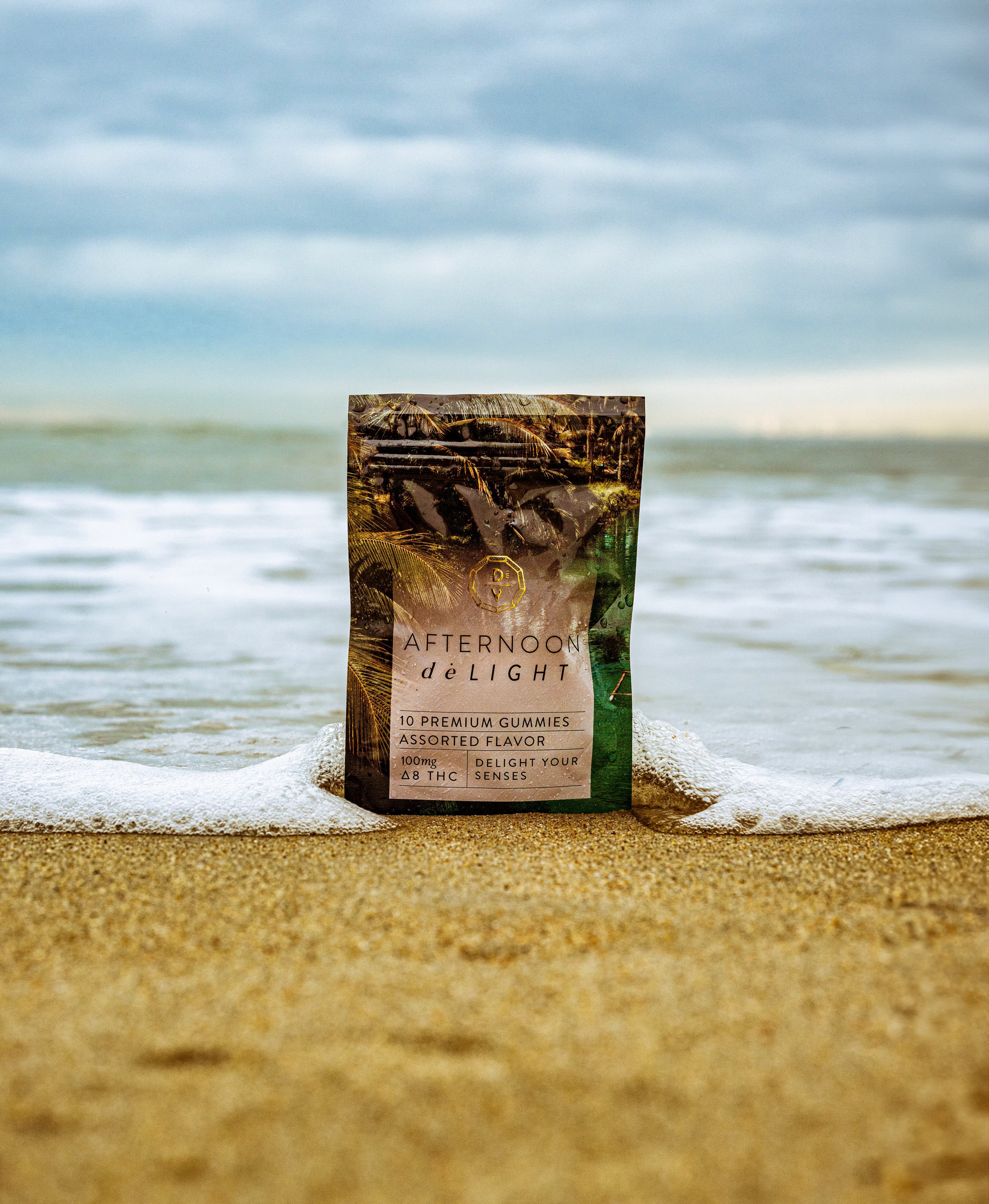 Afternoon deLIGHT ∆8 Gummies pack on sandy beach with ocean waves and cloudy sky background.
