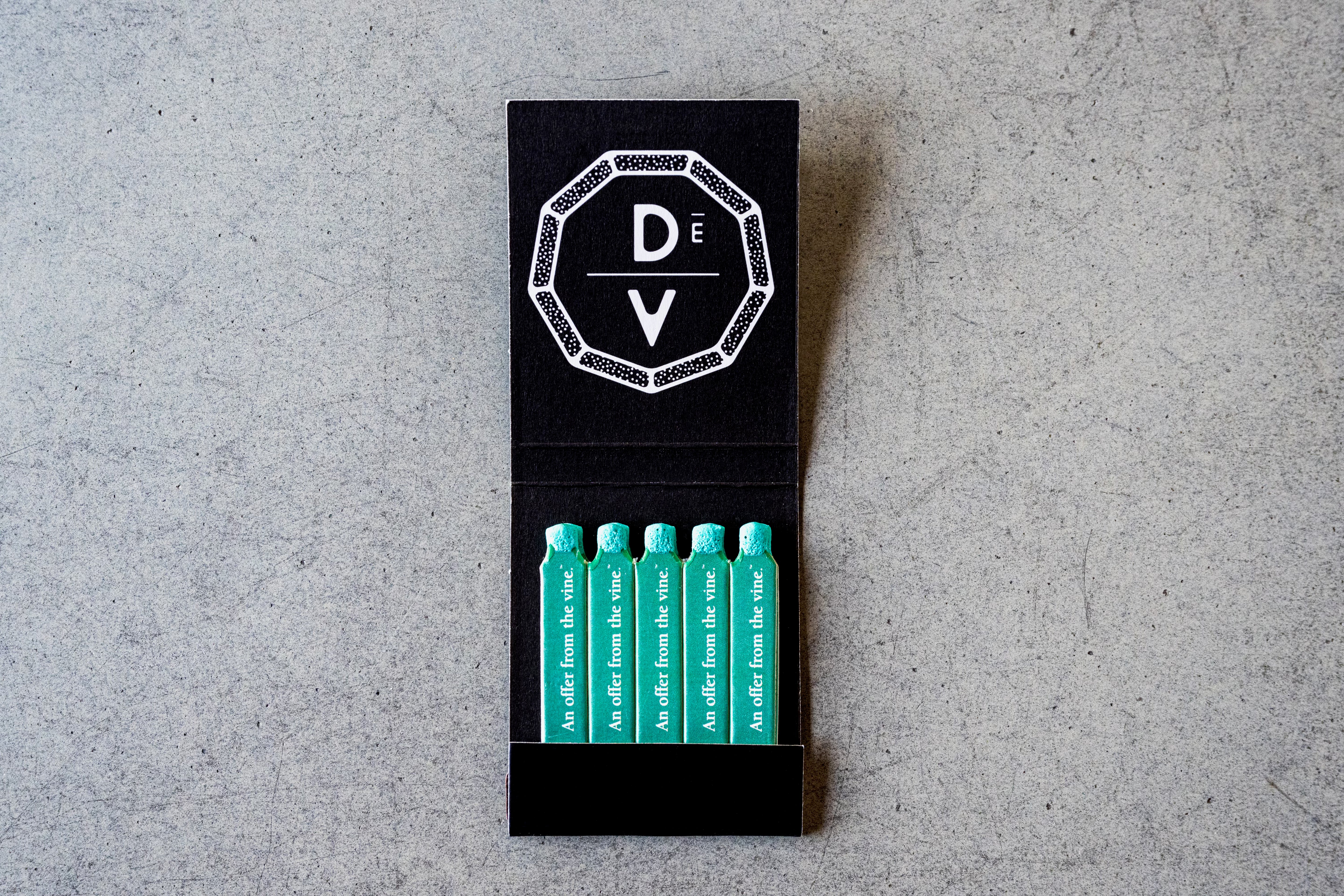 A black matchbook labeled “DÉV” with a white geometric logo, from DeVINE Wellness, opens to reveal five teal deVINE Matches—each printed with I am different from the others—for an elevated smoking experience.