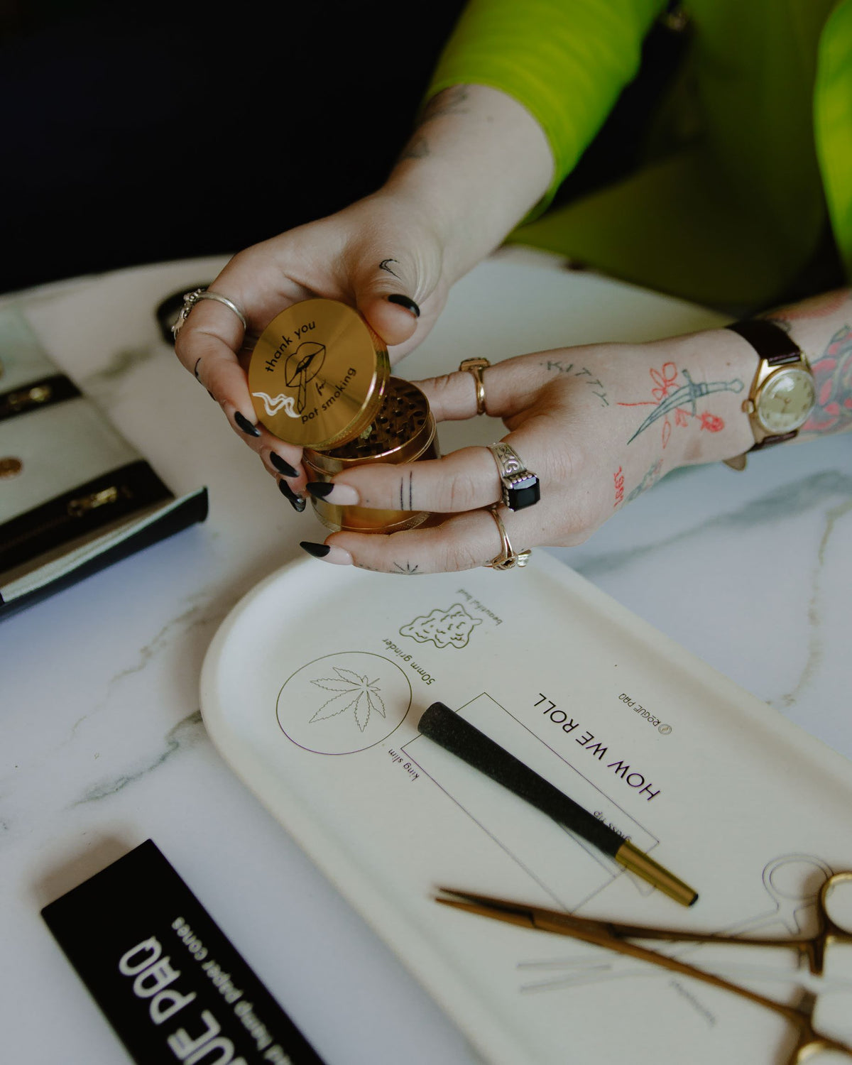 A tattooed, ringed hand holds the open Rogue Paq Thank You For P0t Smoking Grinder with diamond teeth above a HOW WE ROLL tray, amid illustrations, a rolling paper, and smoking accessories on a white surface.