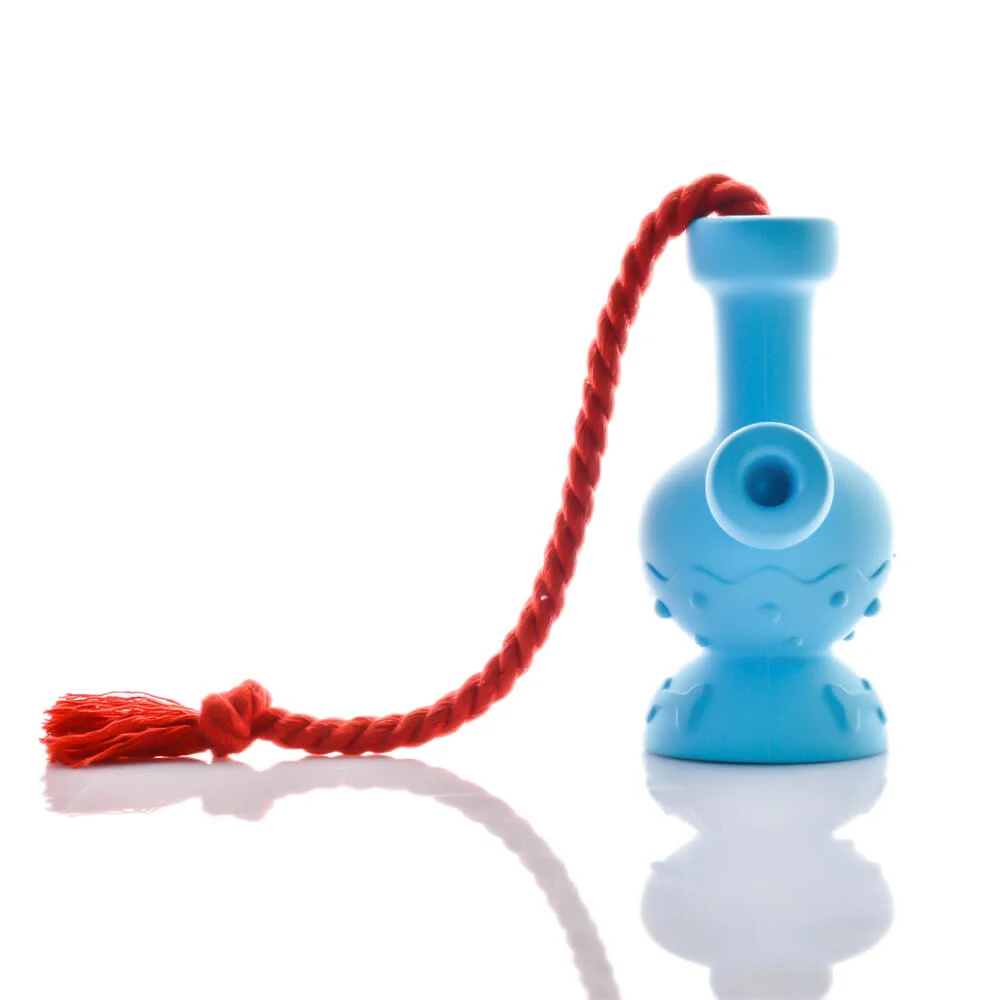 The Tug & Toke by Puff Palz is a small blue natural rubber dog toy shaped like a vase, with a textured base, round side hole, and thick red rope ending in a tassel, shown on a white reflective surface.