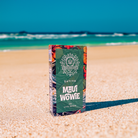 A vibrant DeVINE Destinations Live Resin THC Disposable by DeVINE Wellness stands upright on a sandy beach with clear blue sky and ocean waves, evoking travel vibes and premium cannabis relaxation.
