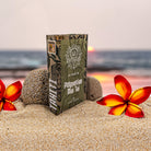 A deVINE Destinations Live Resin THC Disposable by DeVINE Wellness rests on a sandy beach with two red and yellow flowers and smooth stones at sunset—inviting you to a travel-inspired vaping experience.