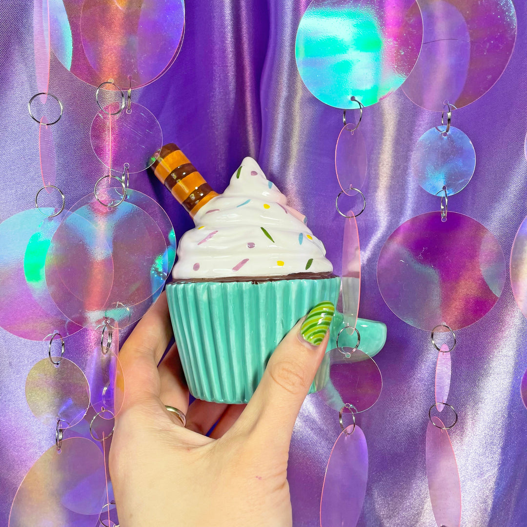 A hand with green, wavy nails holds the Fashioncraft Cupcake Pipe, a turquoise mug-shaped pipe with faux whipped cream, sprinkles, and a chocolate wafer, set against an iridescent sequin background.