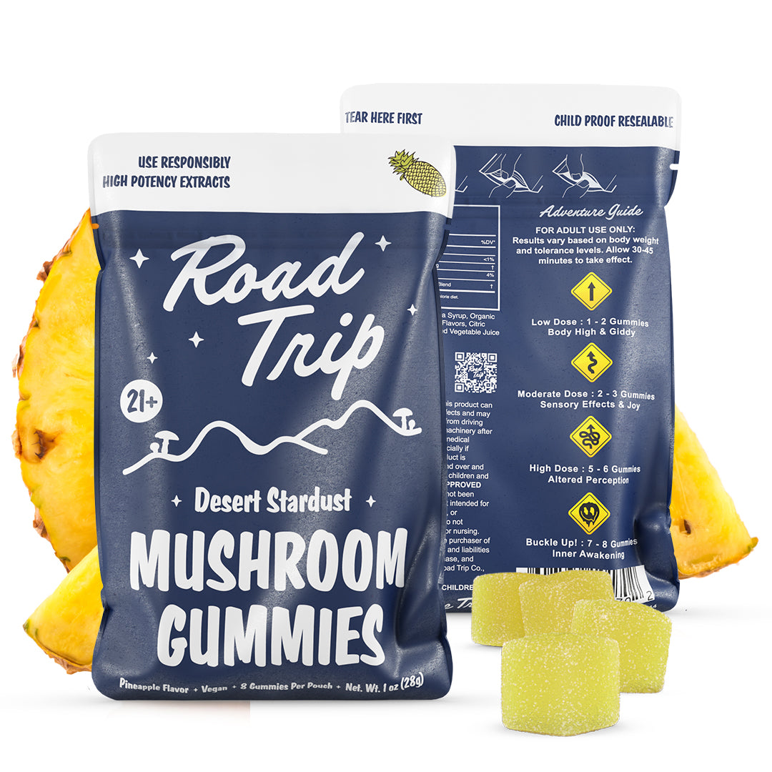 Two navy blue bags of Road Trip Mushroom Gummies from Road Trip are shown with pineapple slices and three yellow, sugar-coated vegan gummies in front. The back bag shows directions and dosage information.