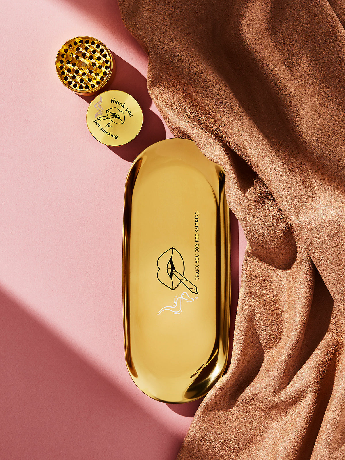 The Rogue Paq Thank You For P0t Smoking Grinder, featuring diamond grind teeth and a built-in kief catcher, pairs stylishly with a minimalist lip tray and soft tan fabric for a chic scene against a pink background.