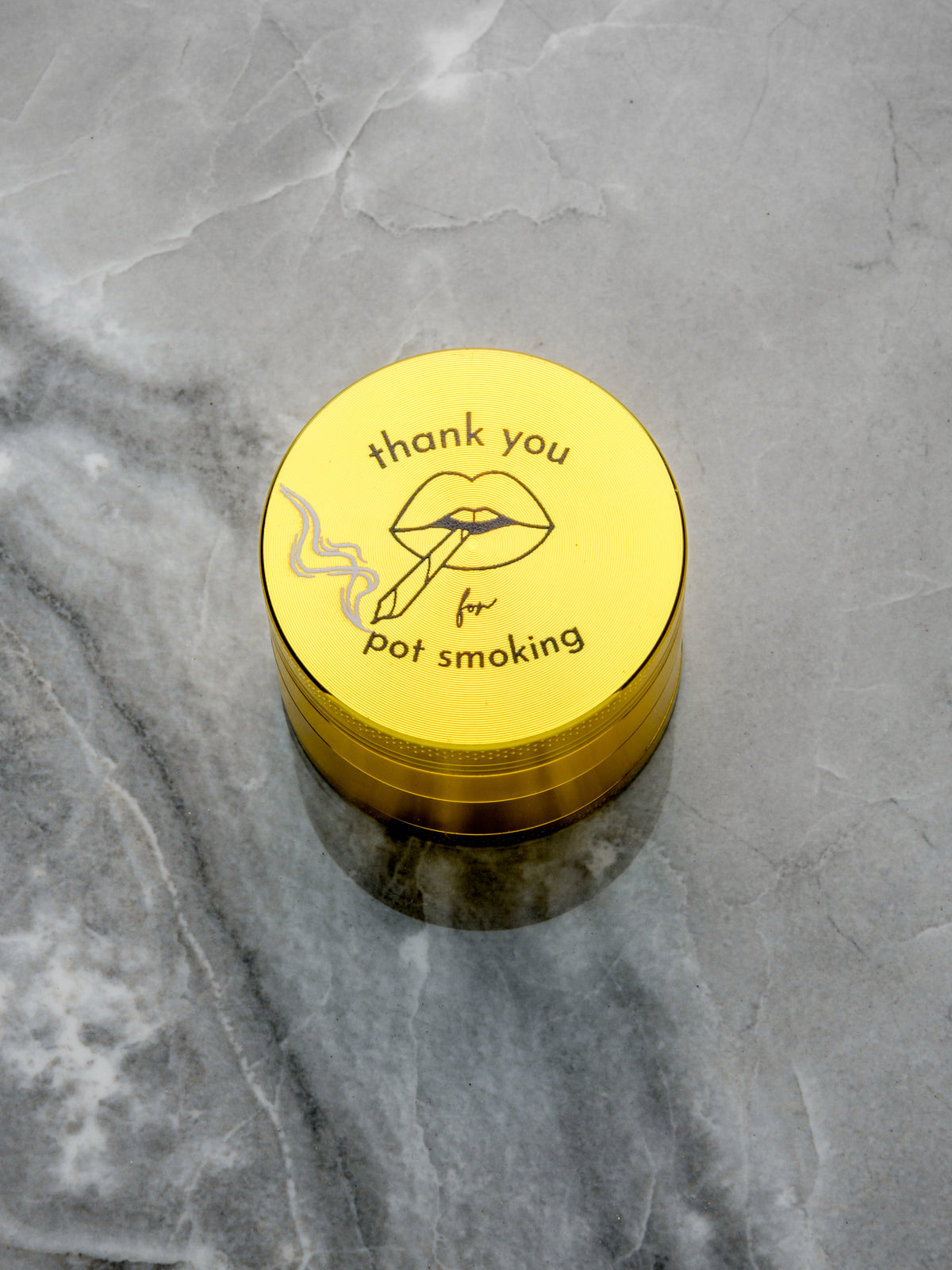 The Rogue Paq Thank You For P0t Smoking Grinder is a round yellow herb grinder with diamond grind teeth, featuring lips smoking and the text thank you for pot smoking, shown on a gray marble surface.