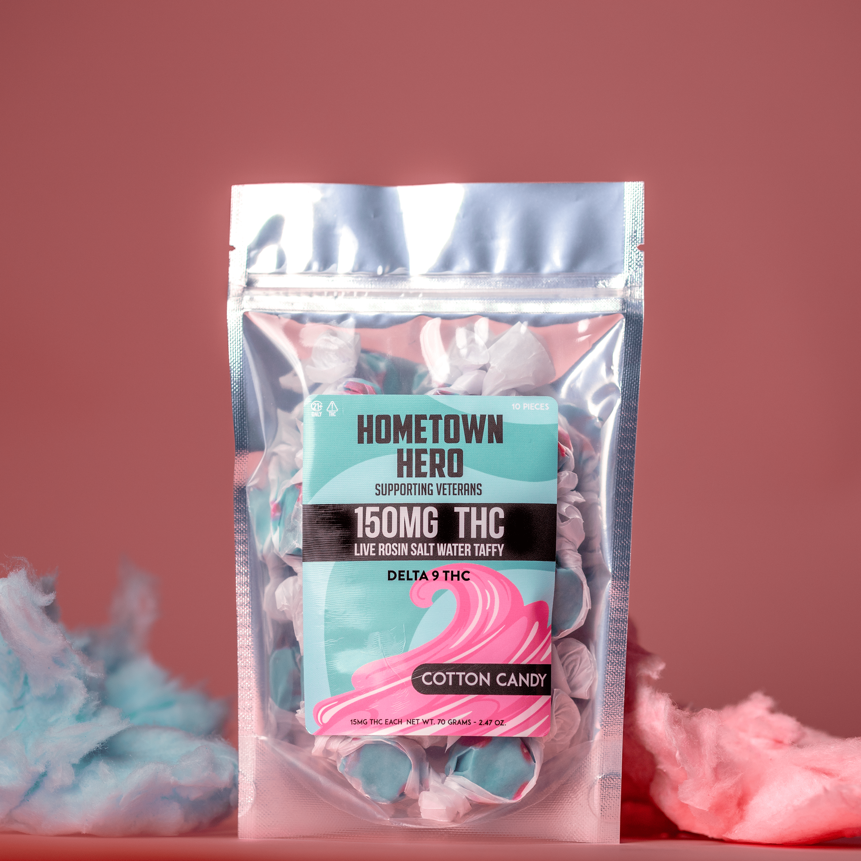 A bag of Hometown Hero Delta-9 Live Rosin Taffy stands against a pink background, surrounded by pink and blue cotton candy, highlighting tasty hemp-derived edibles for those who love live rosin-infused treats.