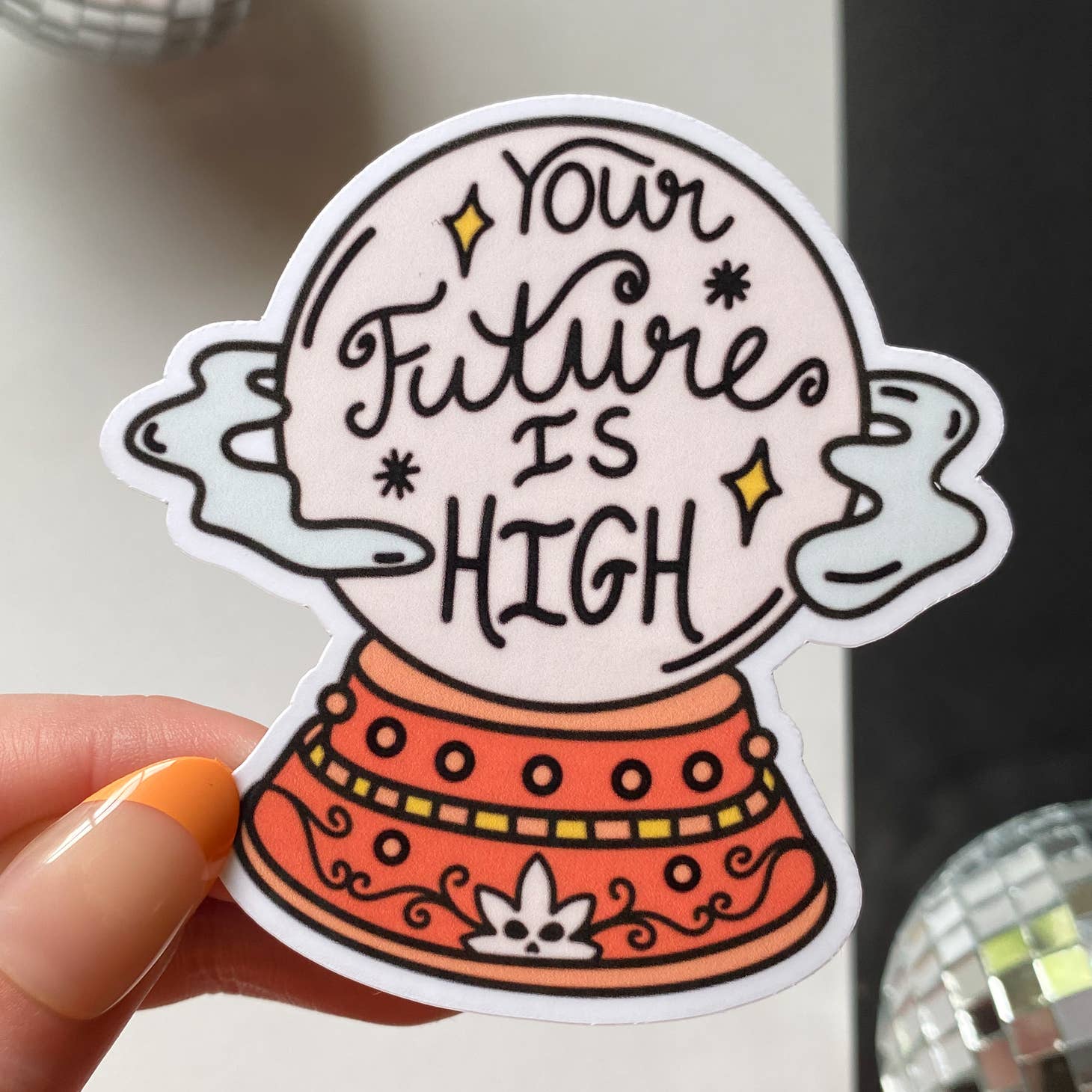 A hand with orange and nude nails holds Han Made’s “Your Future Is High Sticker,” featuring a crystal ball, stars, and a red base—perfect as a waterproof laptop sticker for daily inspiration.