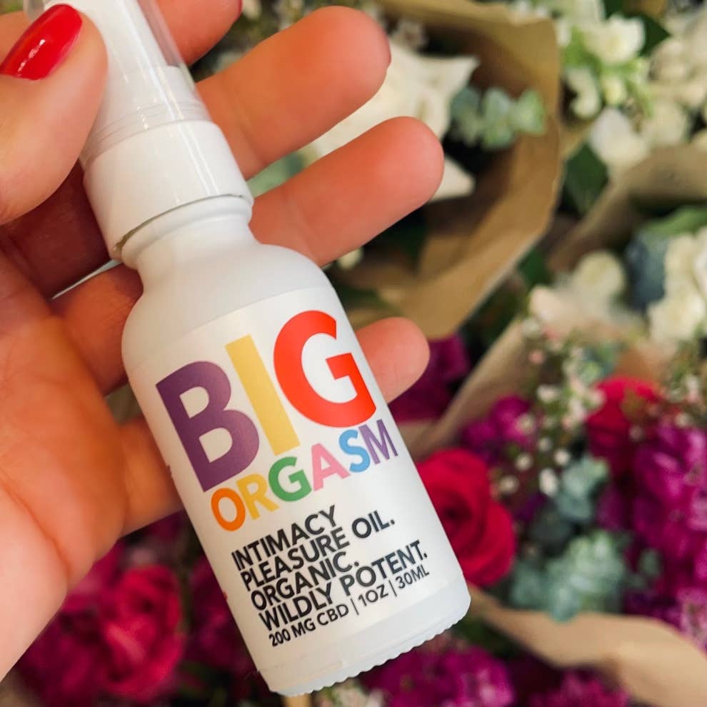 A hand with red-painted nails holds a white bottle labeled Big Orgasm Oil by Eat Gold Organics, featuring colorful text. This organic intimacy oil with damiana leaf is set against a blurred background of assorted flowers.