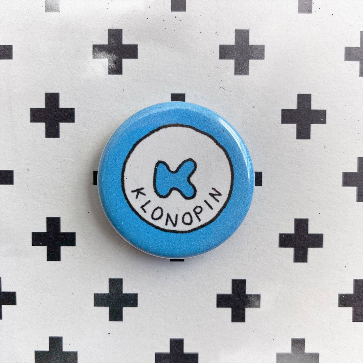 A round blue Pill Pin by Chateau Blanche, featuring a white circle with a stylized K and KLONOPIN below it, set against a black plus sign background—a distinctive mental health accessory.