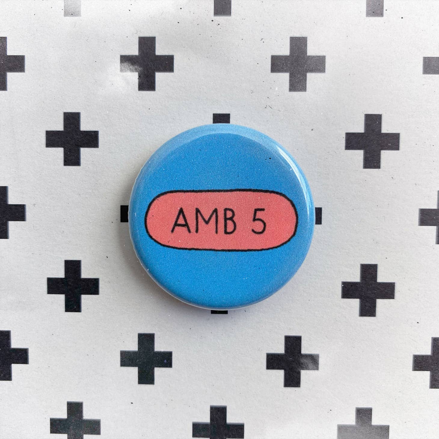 Chateau Blanches Pill Pin—a blue enamel pin with a pink oval center and “AMB 5” text—rests on a white card with a black plus sign pattern, making it a unique accessory for mental health awareness.