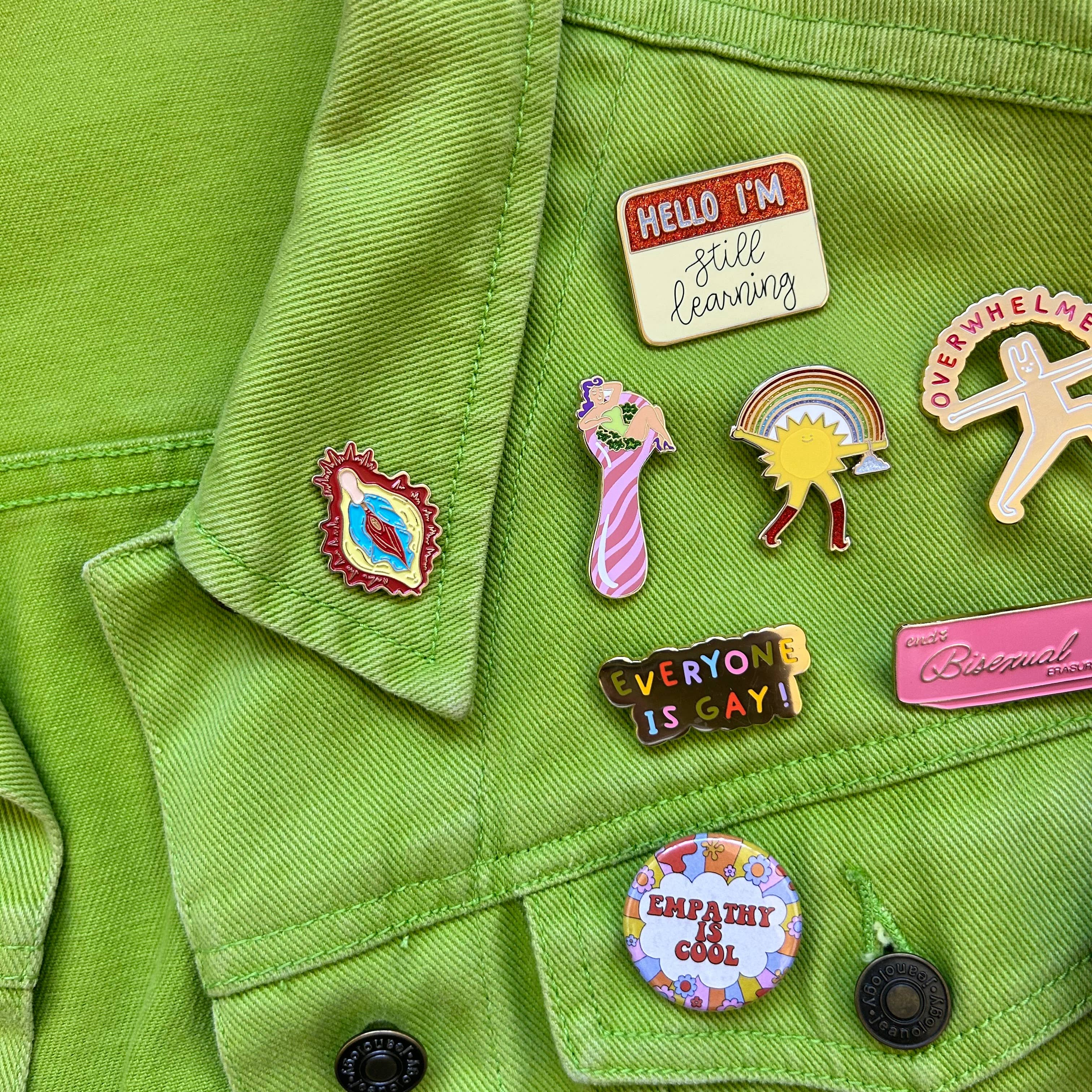 A bright green jacket decorated with colorful enamel pins—glitter, “Hello I’m still learning,” “Everyone is gay!,” “Empathy is cool,” plus rainbows, stars, and The Peach Fuzzs playful Total Smoke Show Enamel Pin.
