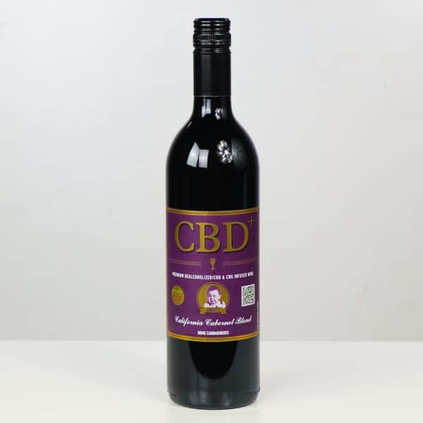 A bottle of Cry Baby Wine’s CBD Cabernet with a purple and gold label, illustrated portrait, and text showing it’s a California Cabernet Blend infused with hemp-derived CBD stands on a white surface against a plain background.