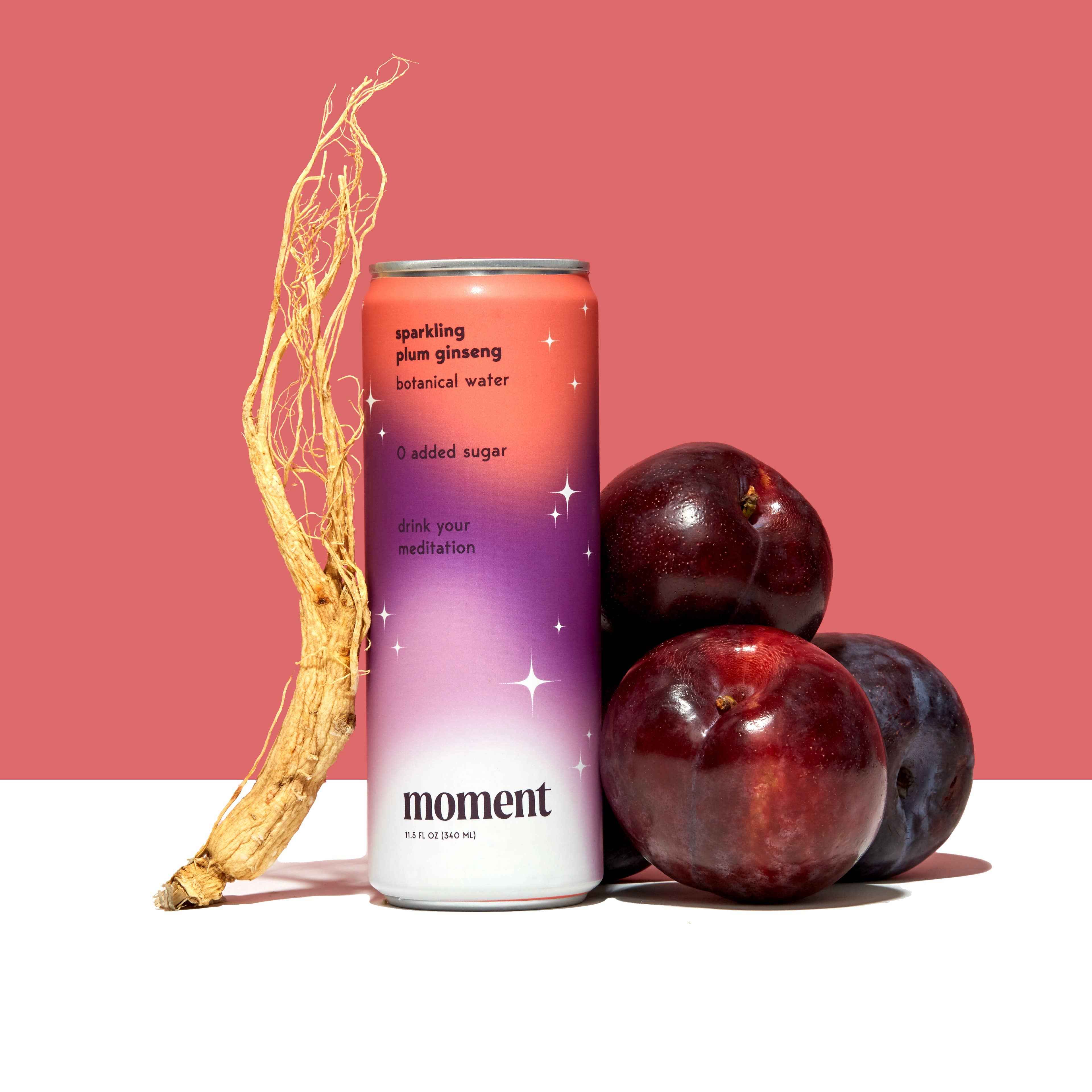 A can of Moment Botanical Beverage by Moment stands upright next to a ginseng root and three dark purple plums, set against a pink and white two-tone background.
