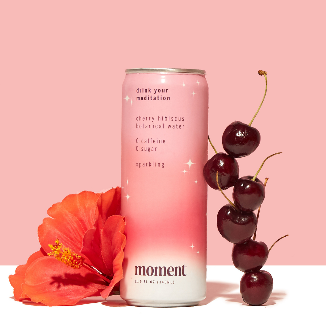 A can of Moment Botanical Beverage from Moment stands upright against a pink background, with a red hibiscus flower to the left and fresh cherries to the right.