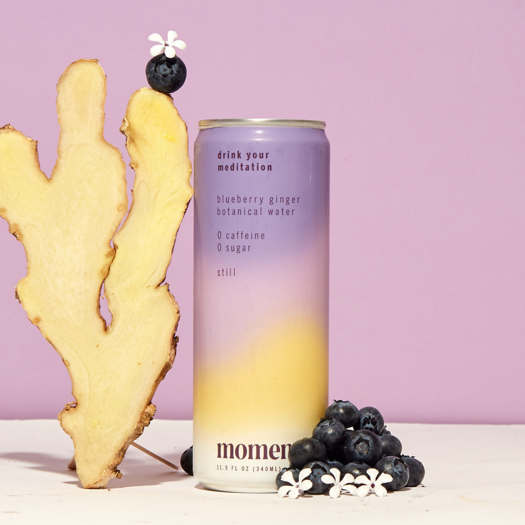 A can of Moment Botanical Beverage by Moment sits next to a ginger slice, blueberries, and white flowers on a pastel purple background.