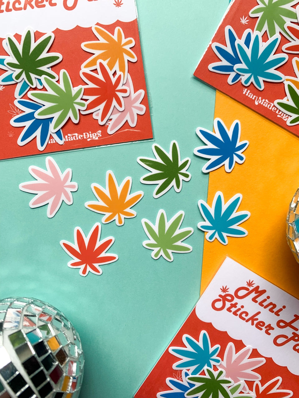 Bright Mini Rainbow Weed Leaf stickers by Han Made are scattered on a teal and yellow background, alongside red Mini Rainbow Weed Leaf Sticker Pack packaging and disco balls—ideal for fans of bold, fun vinyl stickers.
