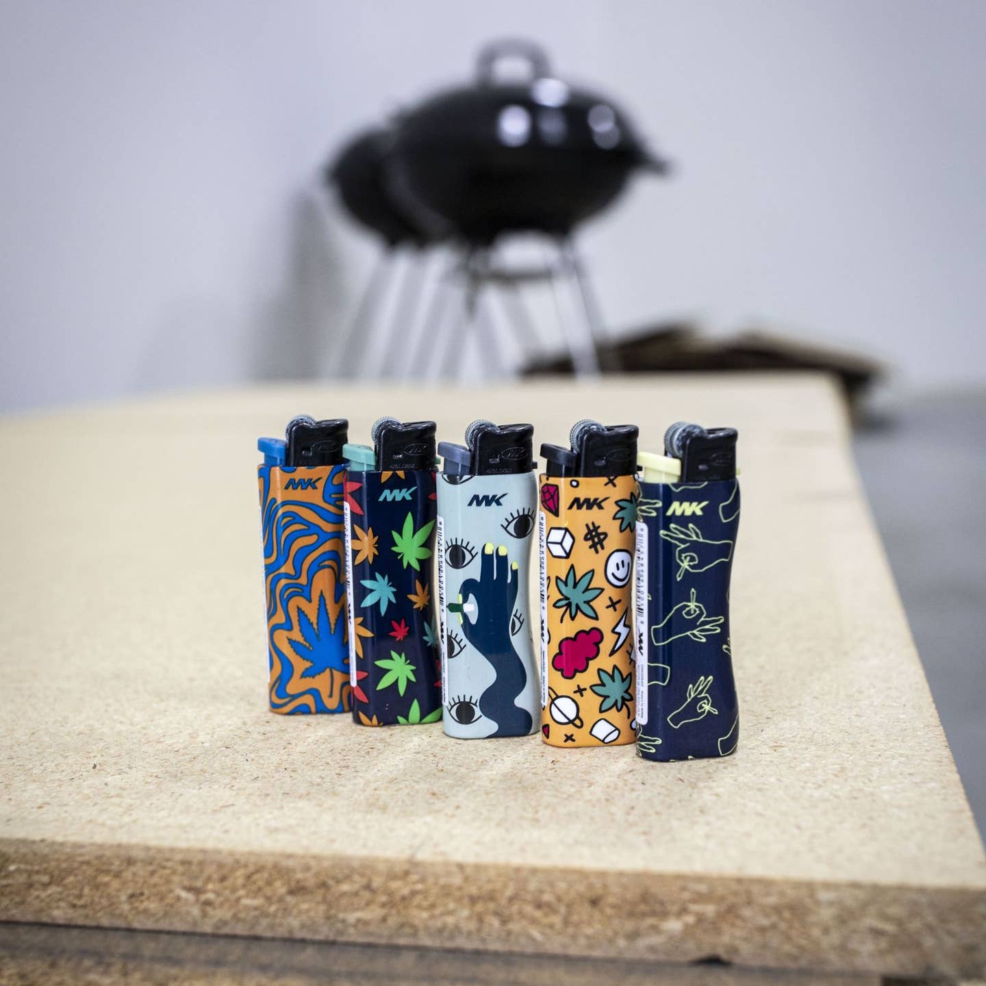 Five vibrant MK Lighter Company Flint Strike Refillable Lighters featuring artistic designs stand upright on wood. Each lighter includes a replaceable flint for long-lasting use, with a blurred grill and outdoor background.