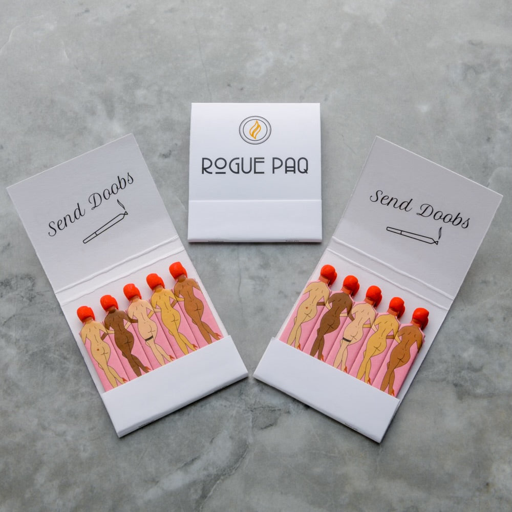 Two open Send Doobs Matches from Rogue Paq, featuring illustrated nudes and red-tipped matches, plus one closed matchbook with the Rogue Paq logo, sit on a gray surface—a bold, playful take on cannabis accessories.