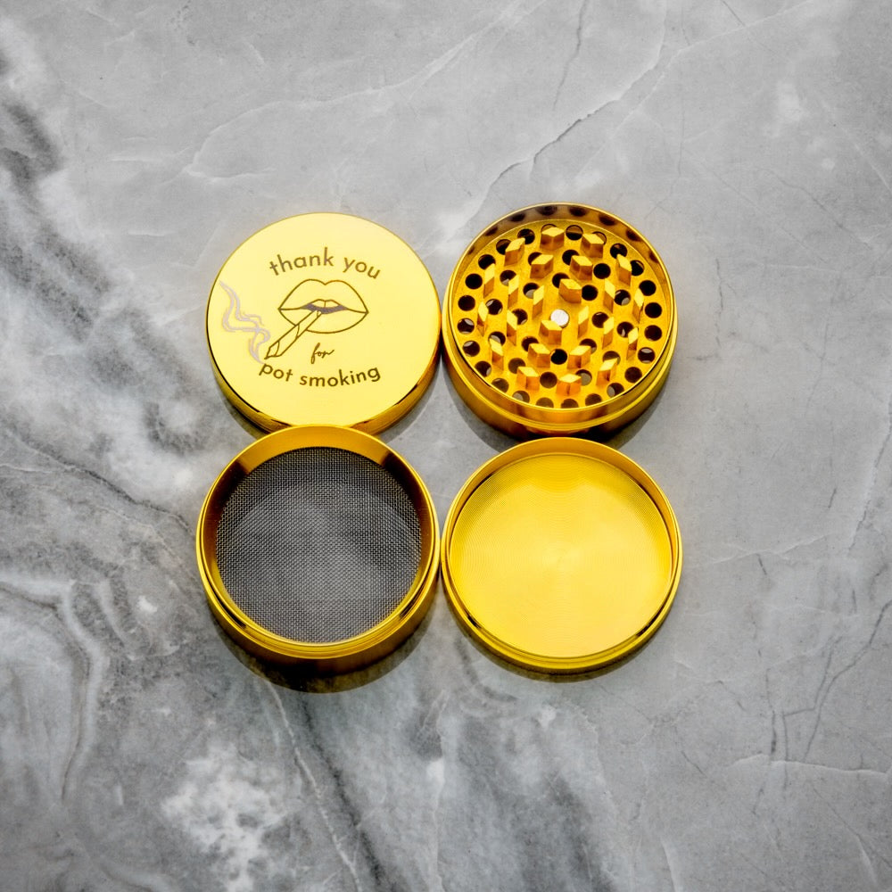 The Rogue Paq Thank You For P0t Smoking Grinder, a gold four-piece herb grinder with diamond grind teeth, sits open on gray marble. Its lid displays lips blowing smoke and the phrase thank you for pot smoking.